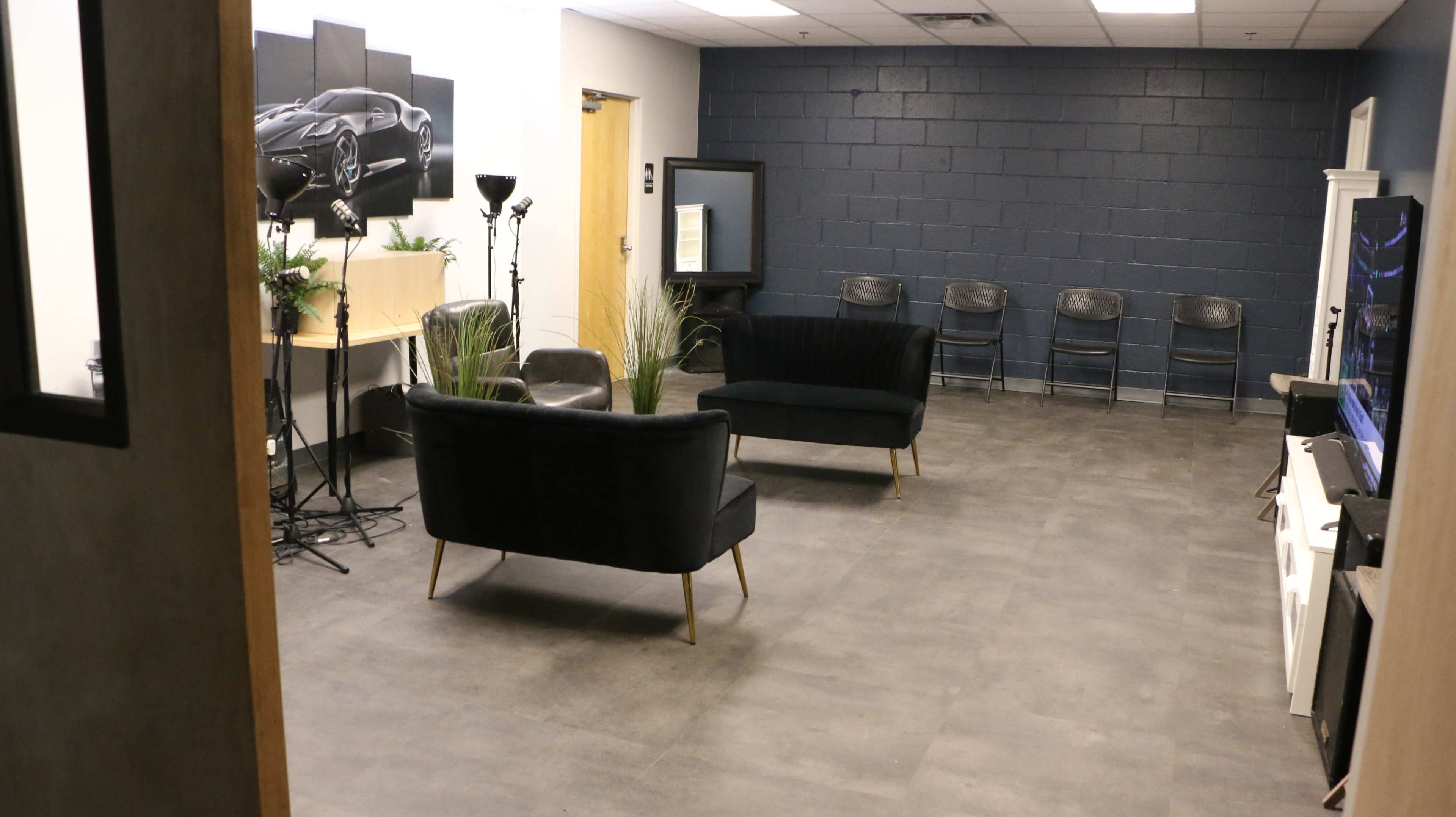 The image shows a waiting room featuring two dark sofas, several chairs, a television, and studio equipment against a blue wall.