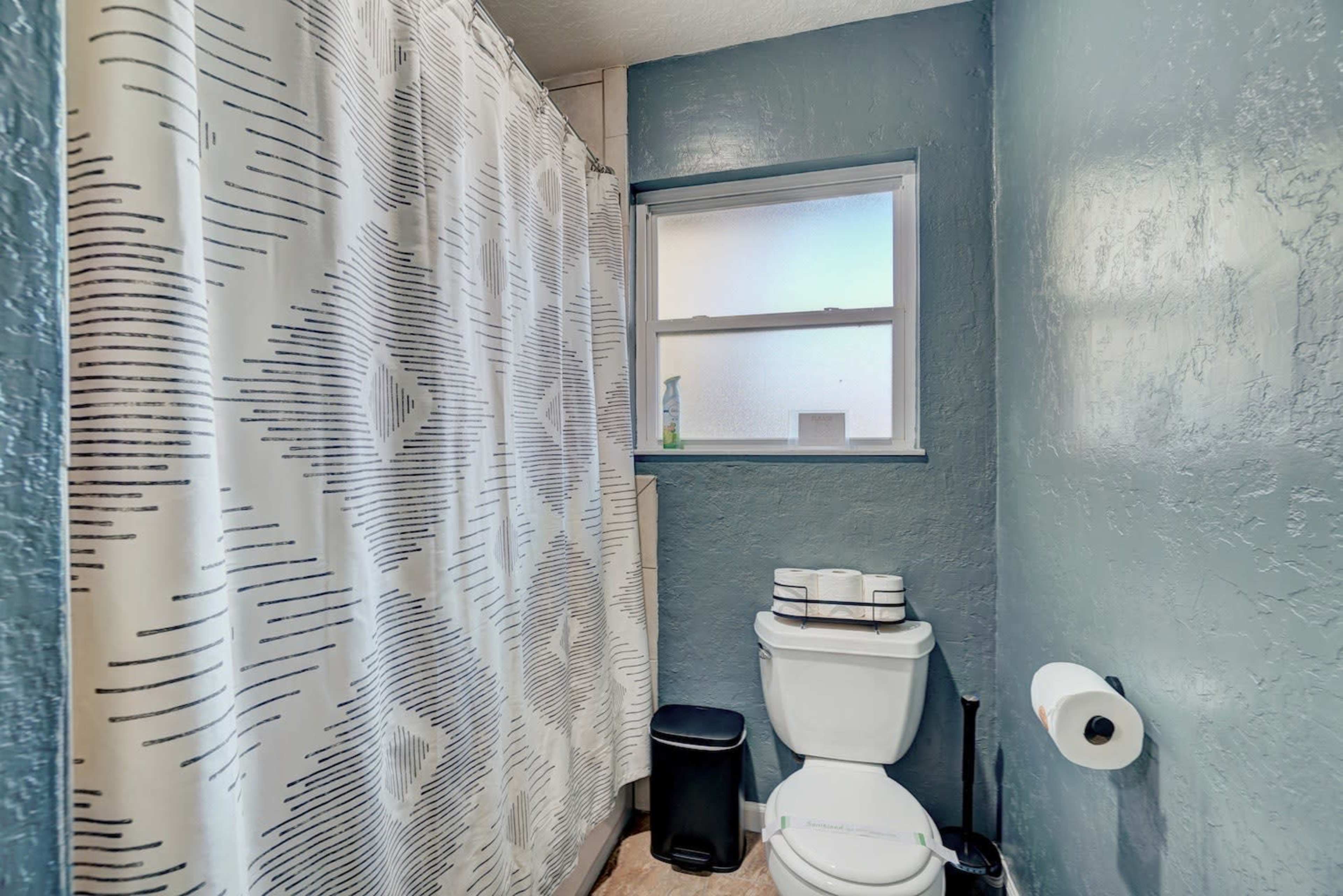 A bathroom features a toilet next to a window, with a patterned shower curtain and a small trash can.