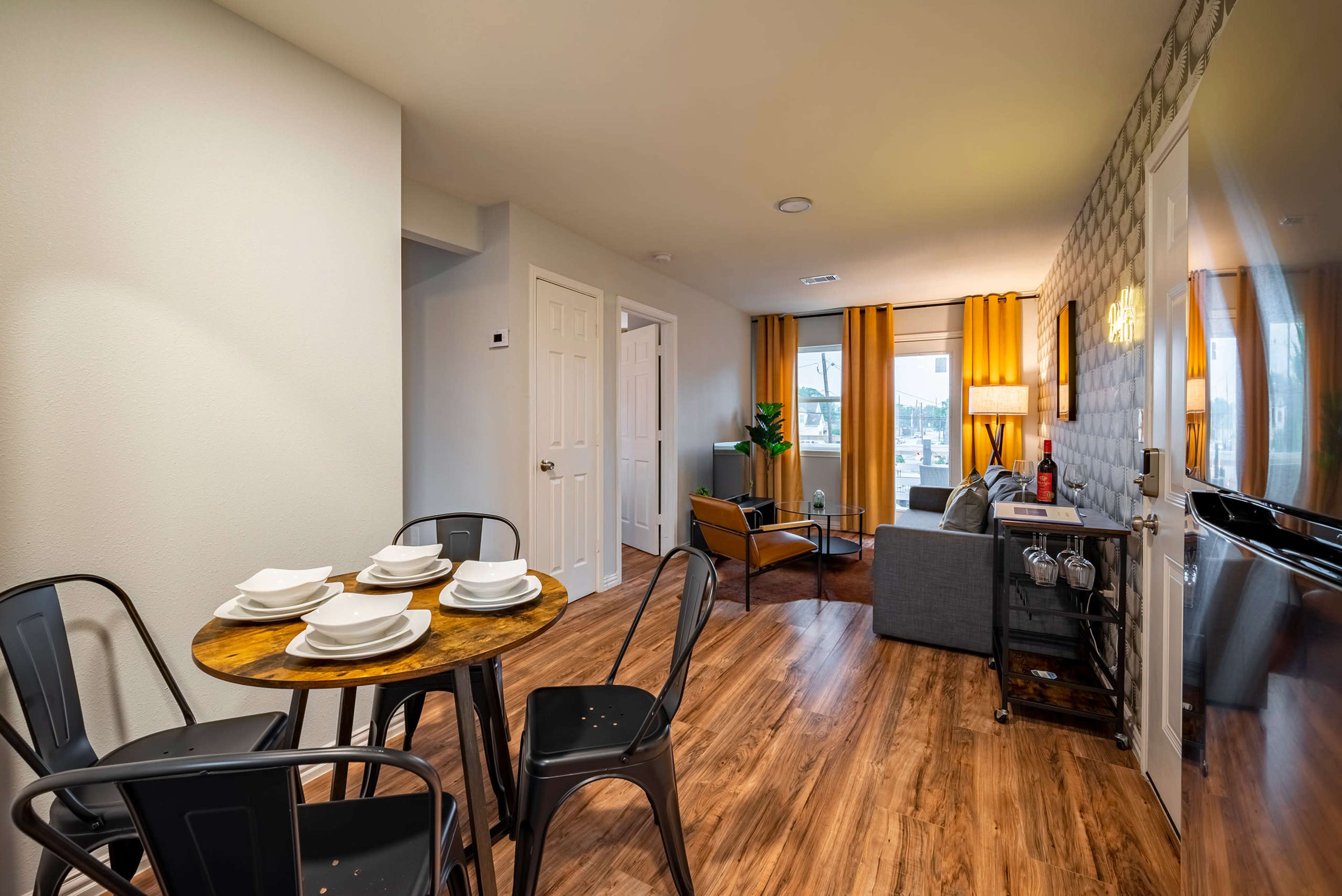 A small dining area with a round wooden table set for four leads into a cozy living space featuring a gray couch and large windows.