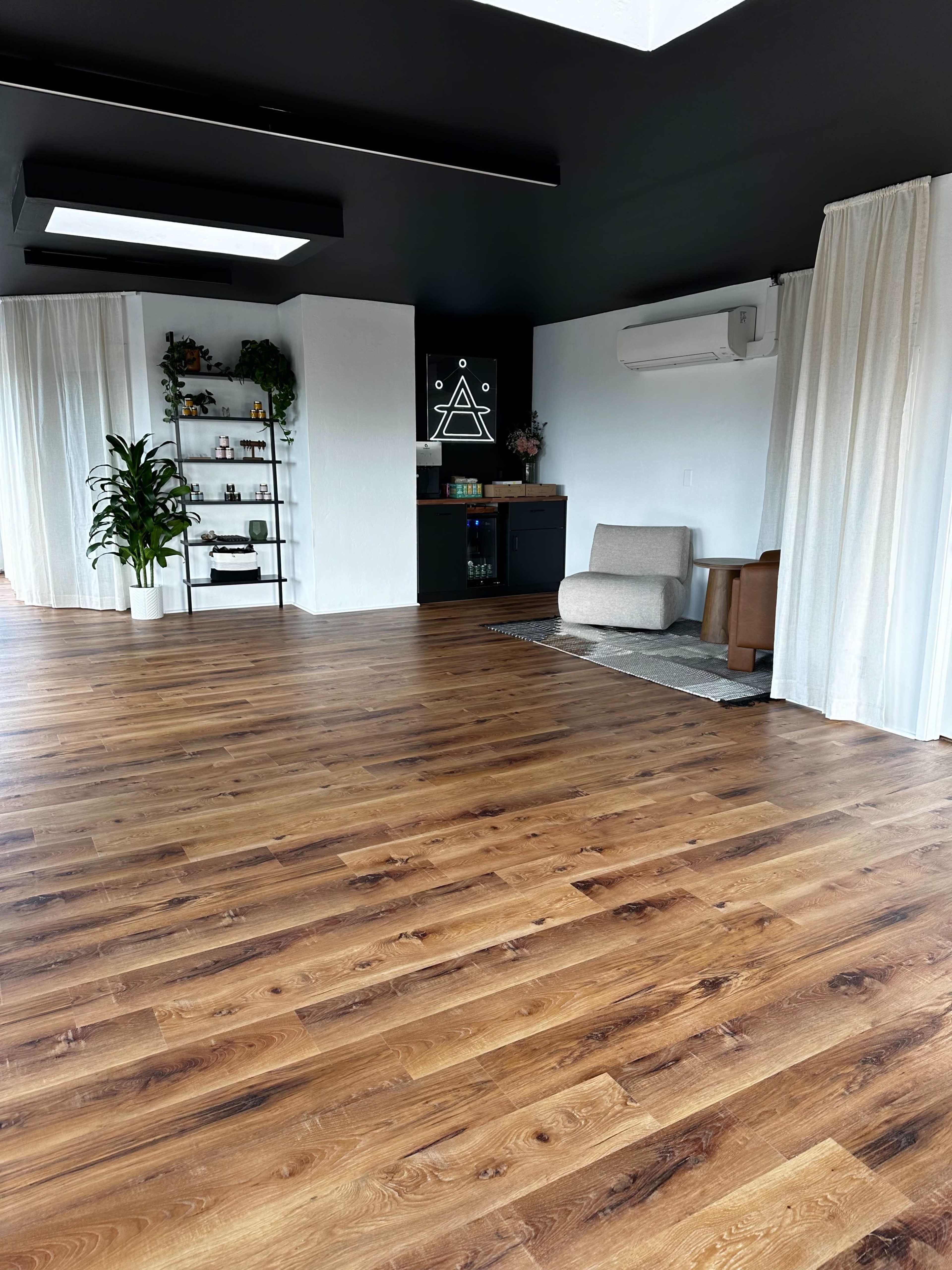 The image shows a spacious interior room with wooden flooring, white curtains, a dark wall featuring a geometric design, and a small bookshelf against one side.
