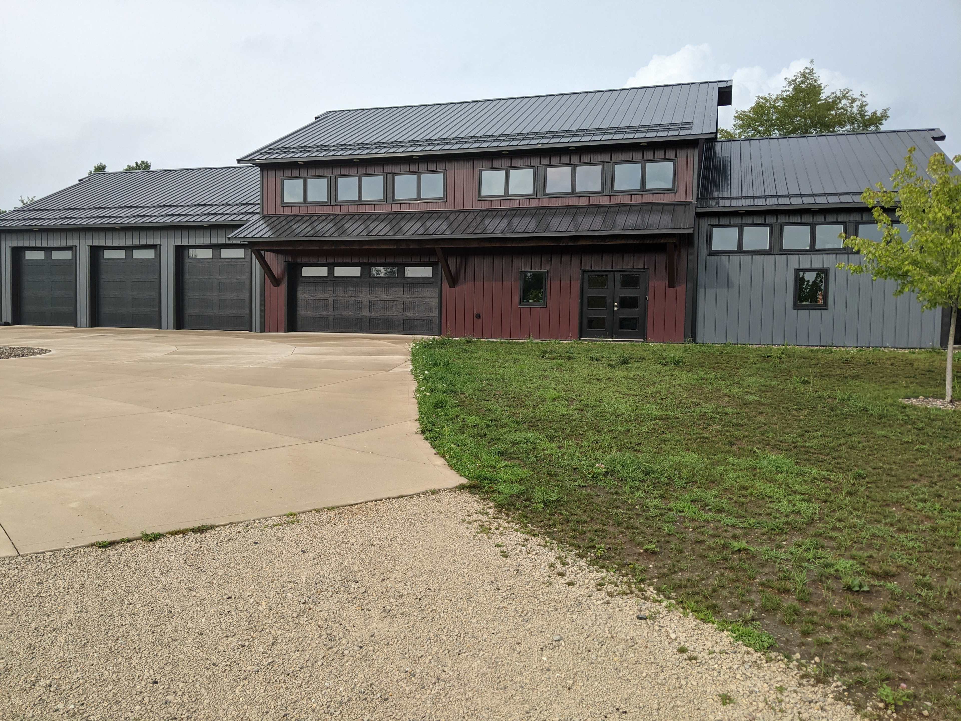 A modern building with a multi-pitched roof features a mix of red and gray metal siding and large windows, set on a concrete driveway surrounded by grass.