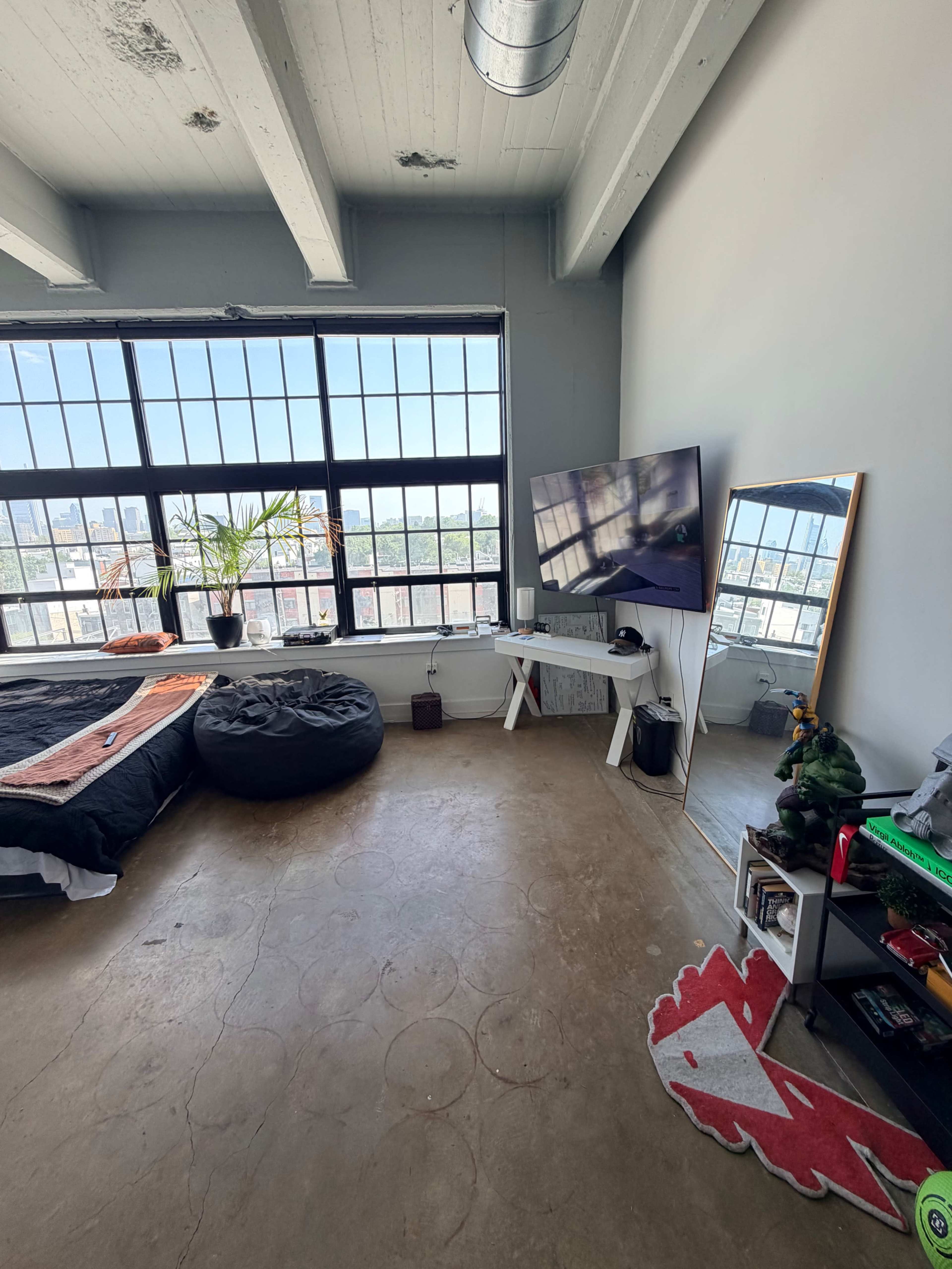 The image shows a modern bedroom with large windows, a television on a stand, a bean bag chair, and minimal furnishings.