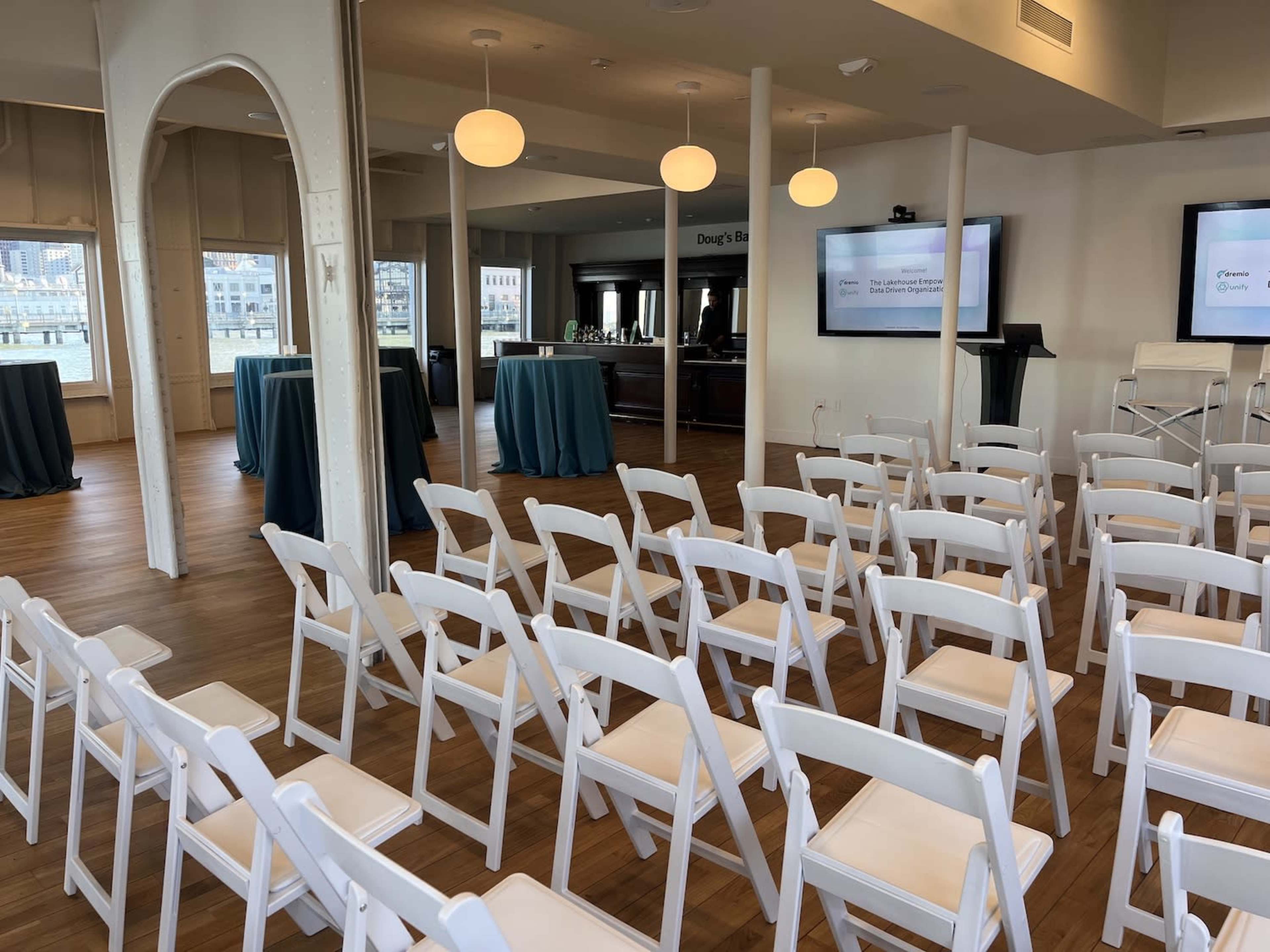A set of white folding chairs arranged in rows facing a presentation area in a well-lit indoor space with tables covered in green cloths.