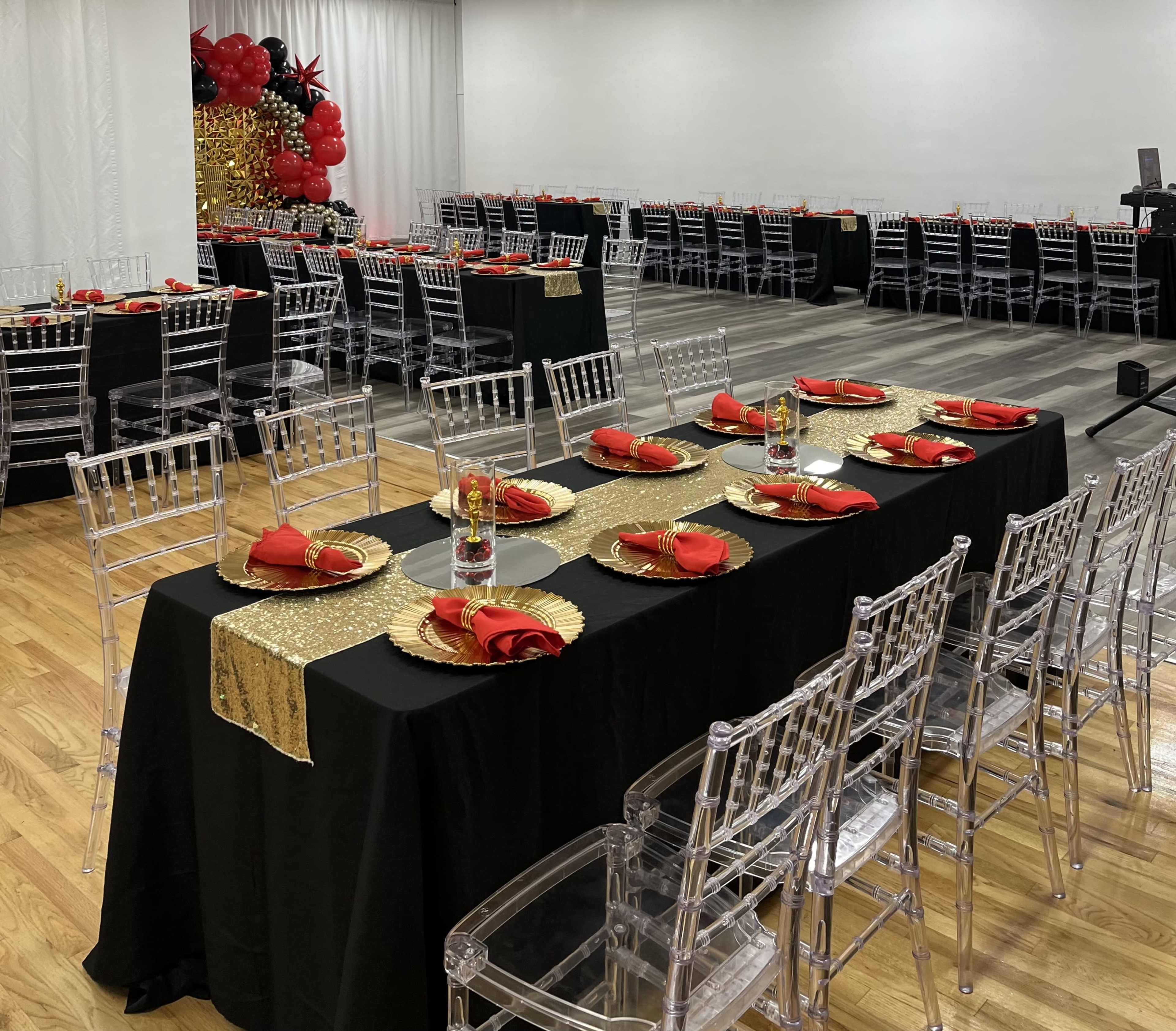 A banquet hall is set up with long tables covered in black tablecloths, featuring red napkins, gold place settings, and clear chairs, with red and black balloon decorations in the background.