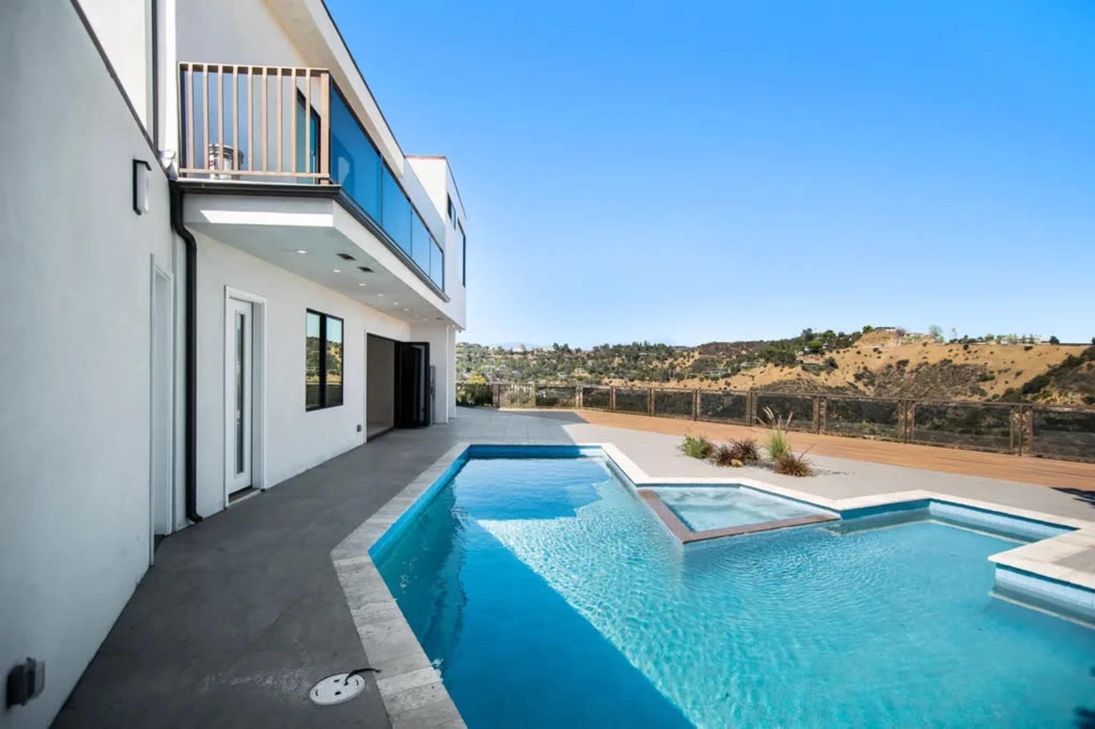 A modern house features a pool with a spa area and a deck overlooking a hillside.