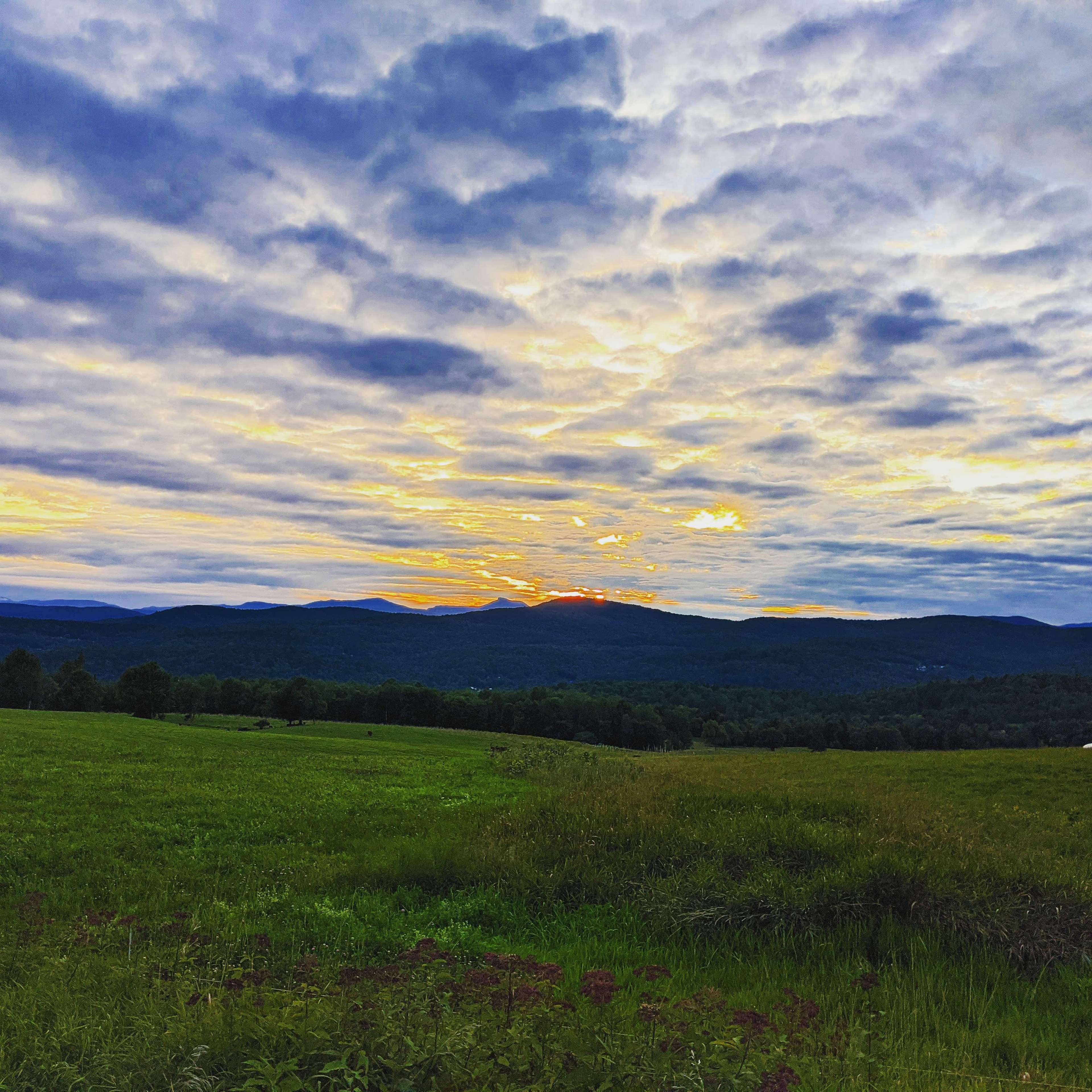 A grassy field in the foreground with mountains and a colorful sunset in the background, under a cloudy sky.