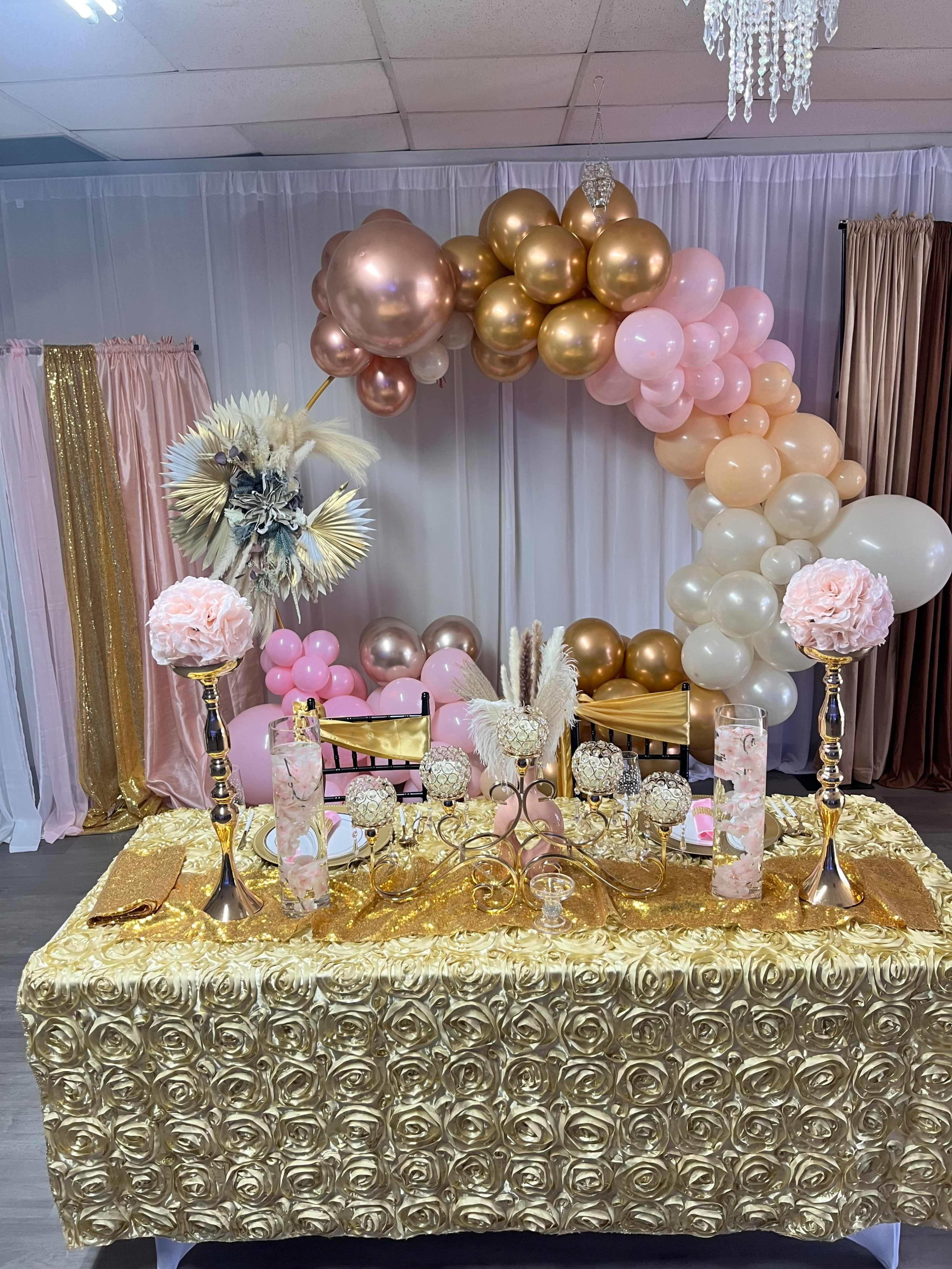 A decorated table with a gold tablecloth adorned with floral arrangements, elegant candles, and a colorful balloon arch in the background.