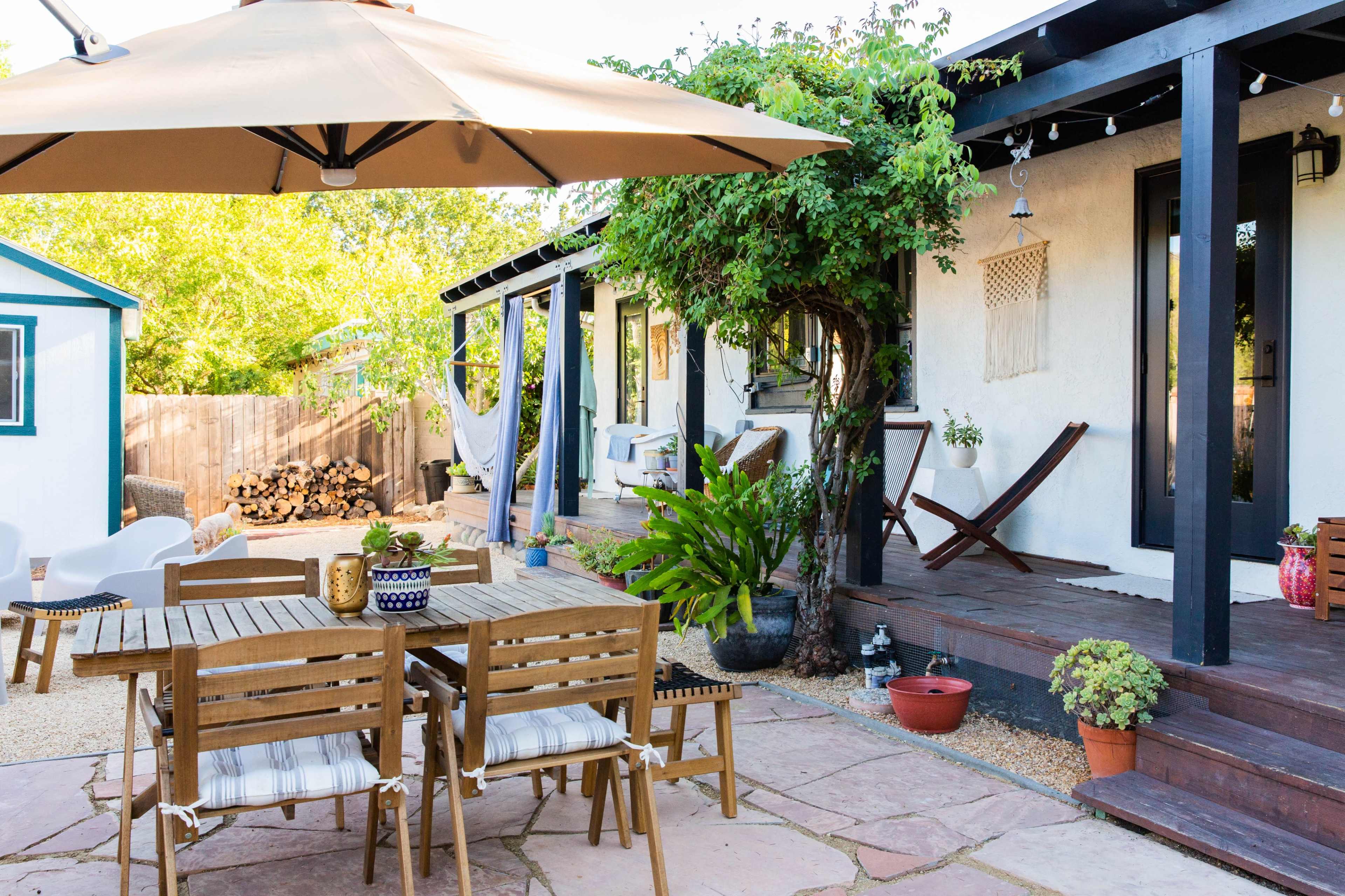 A patio area features a wooden dining table with chairs, shaded by an umbrella, surrounded by potted plants and a firewood stack in the background.