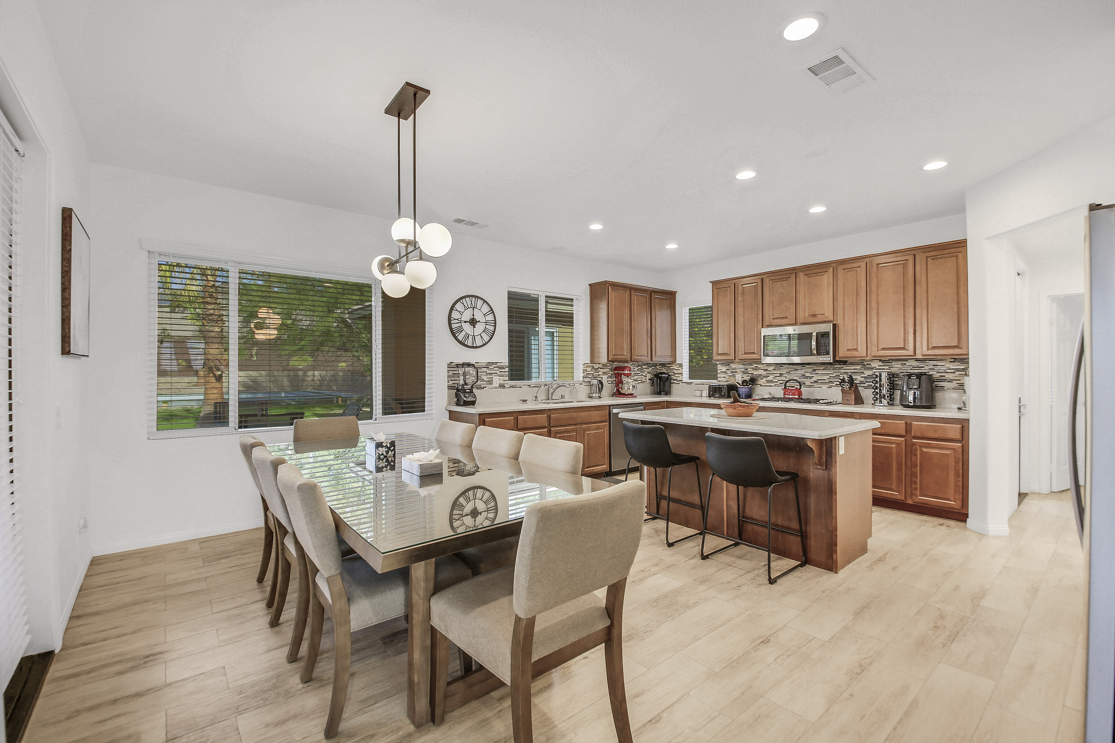 The image shows a modern kitchen and dining area featuring wooden cabinetry, a large dining table with beige chairs, and plenty of natural light from surrounding windows.
