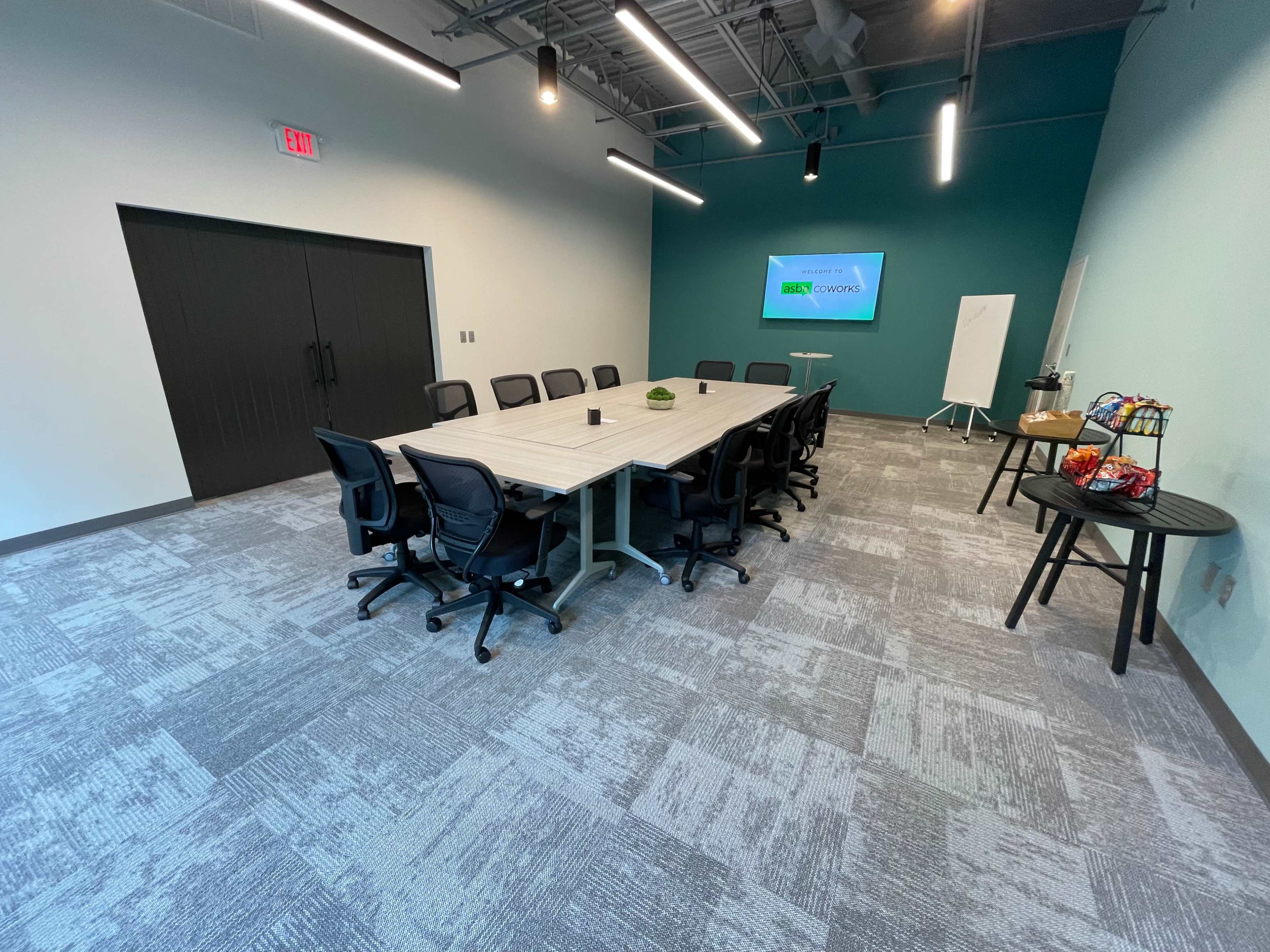 A conference room features a long table surrounded by black chairs, with a large screen and a small refreshment table against a teal wall.