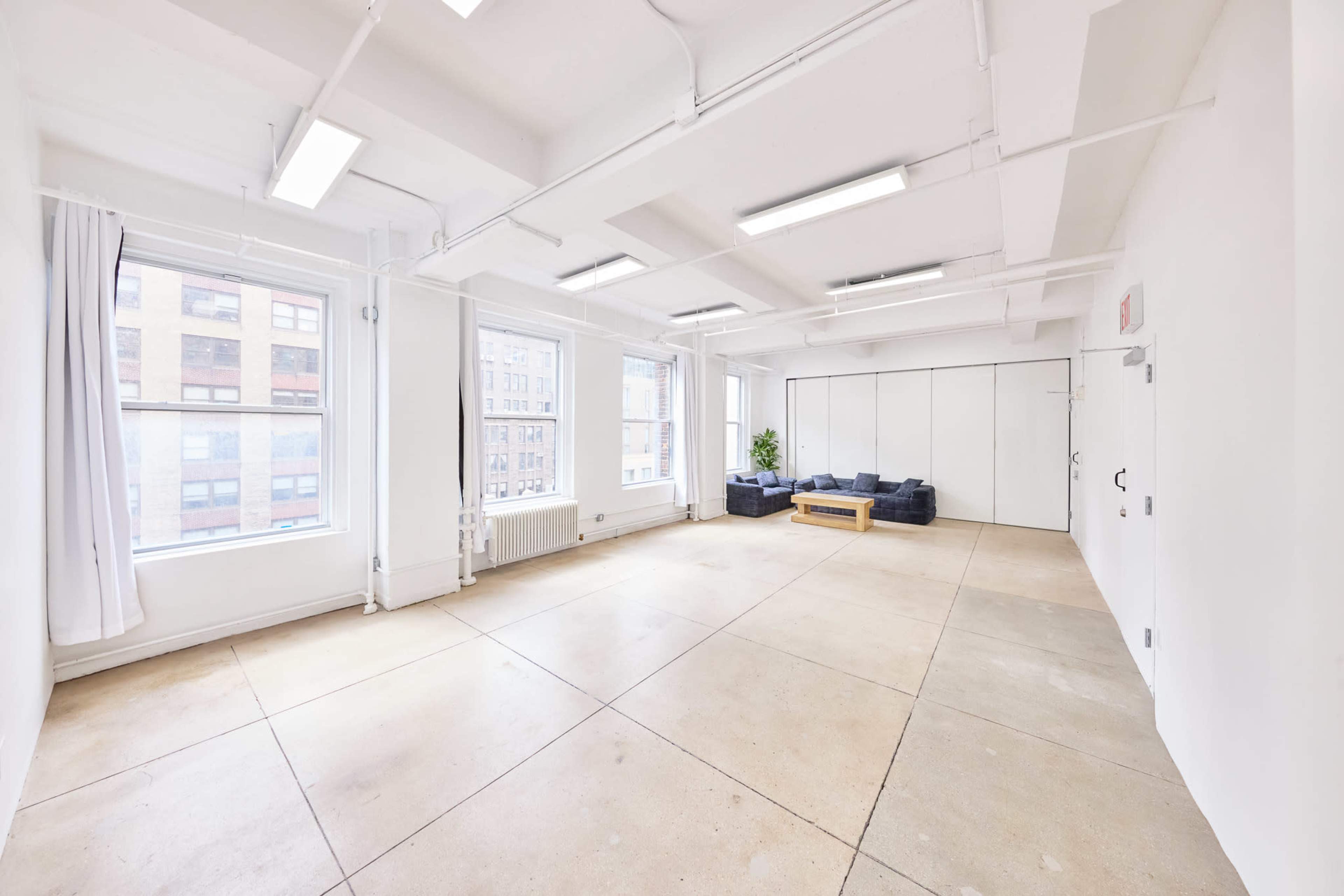 The image shows a spacious, minimalist room with large windows, a seating area featuring a dark sofa, and a small wooden coffee table.