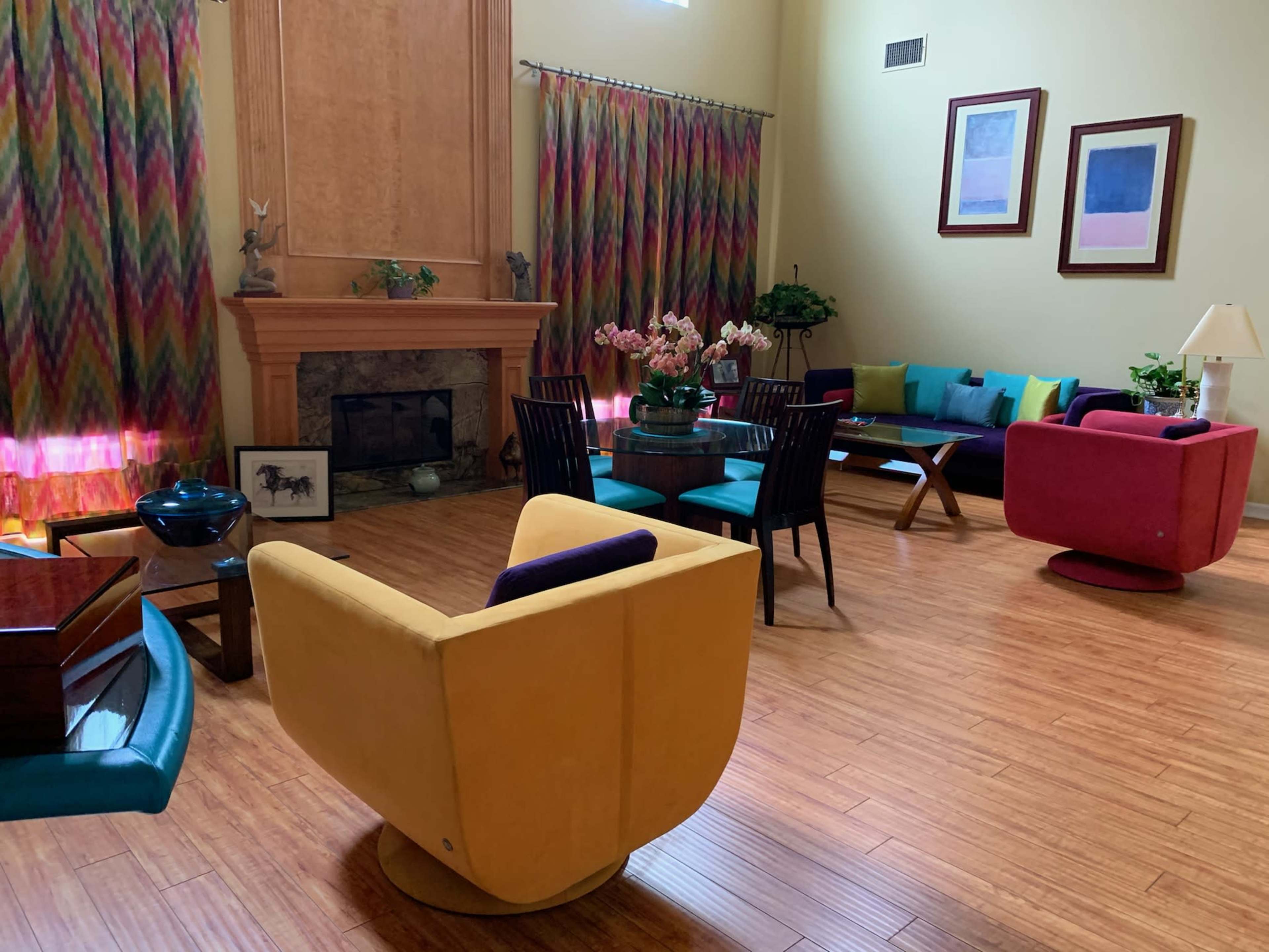 A spacious living room features colorful furniture, a large window with patterned curtains, and a decorative fireplace.