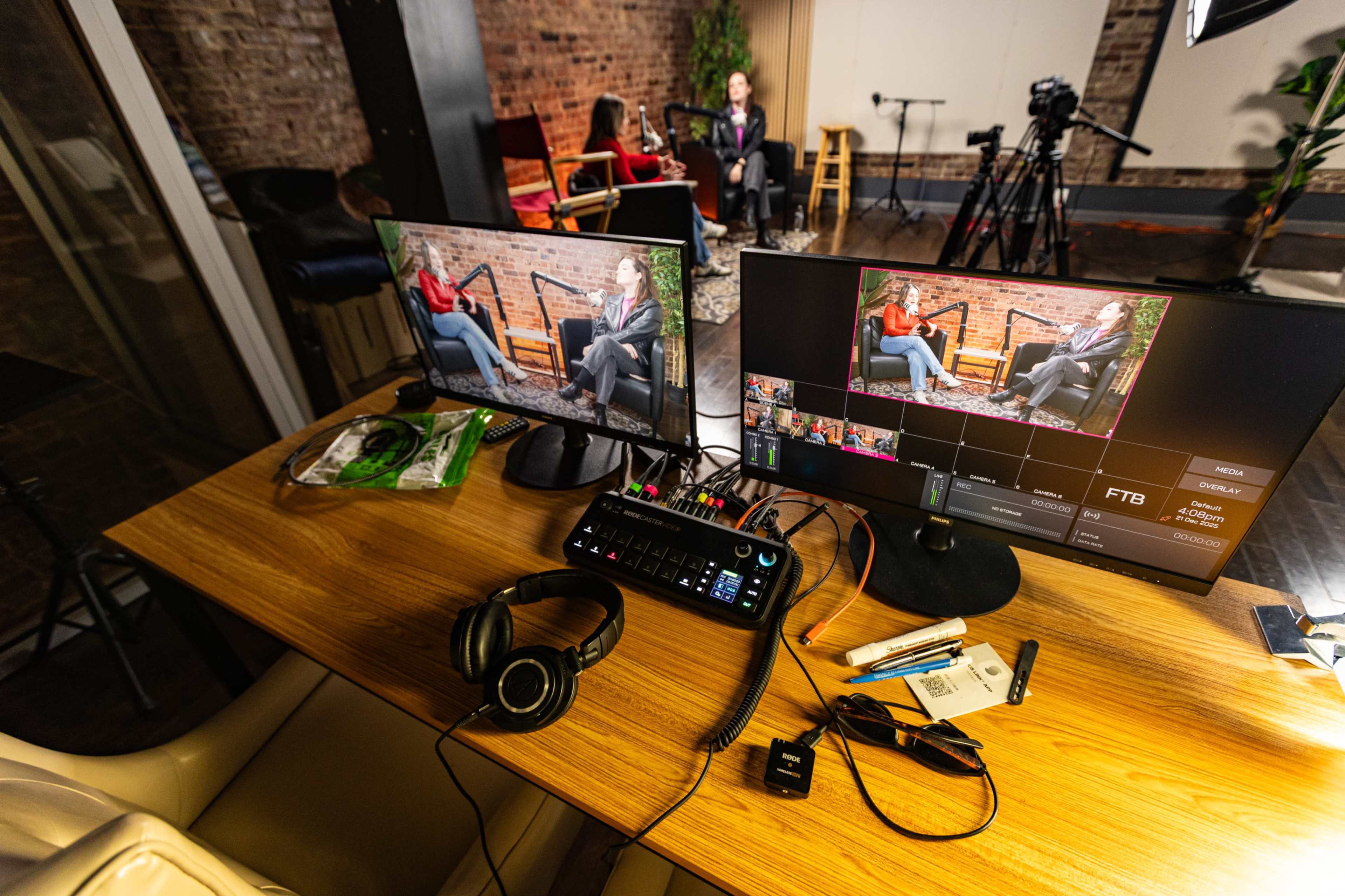 The image shows a video production setup with two monitors displaying interview footage, a control panel, headphones, and various stationery items on a wooden table.