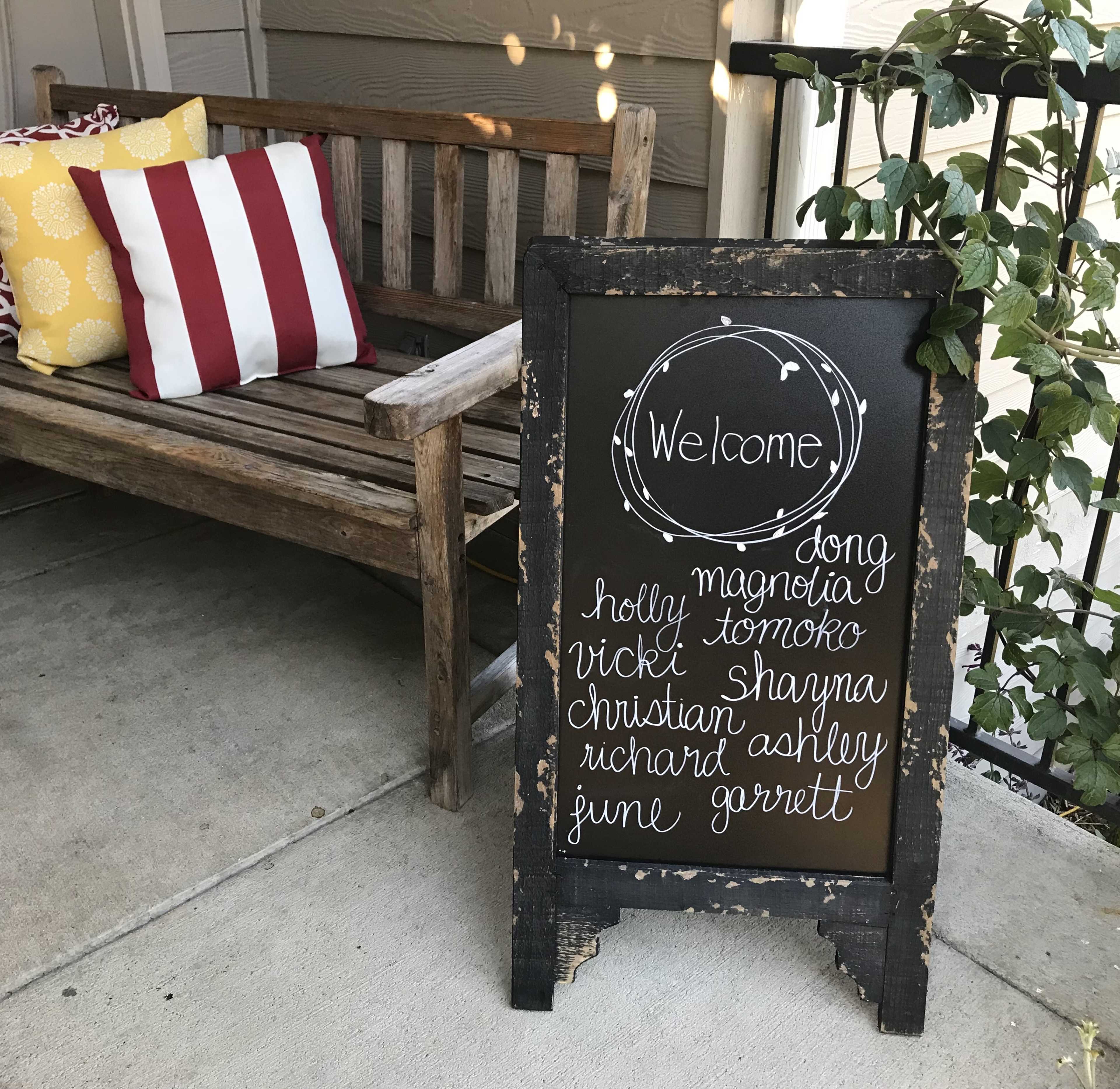 A welcome sign with a list of names stands next to a wooden bench adorned with decorative pillows.