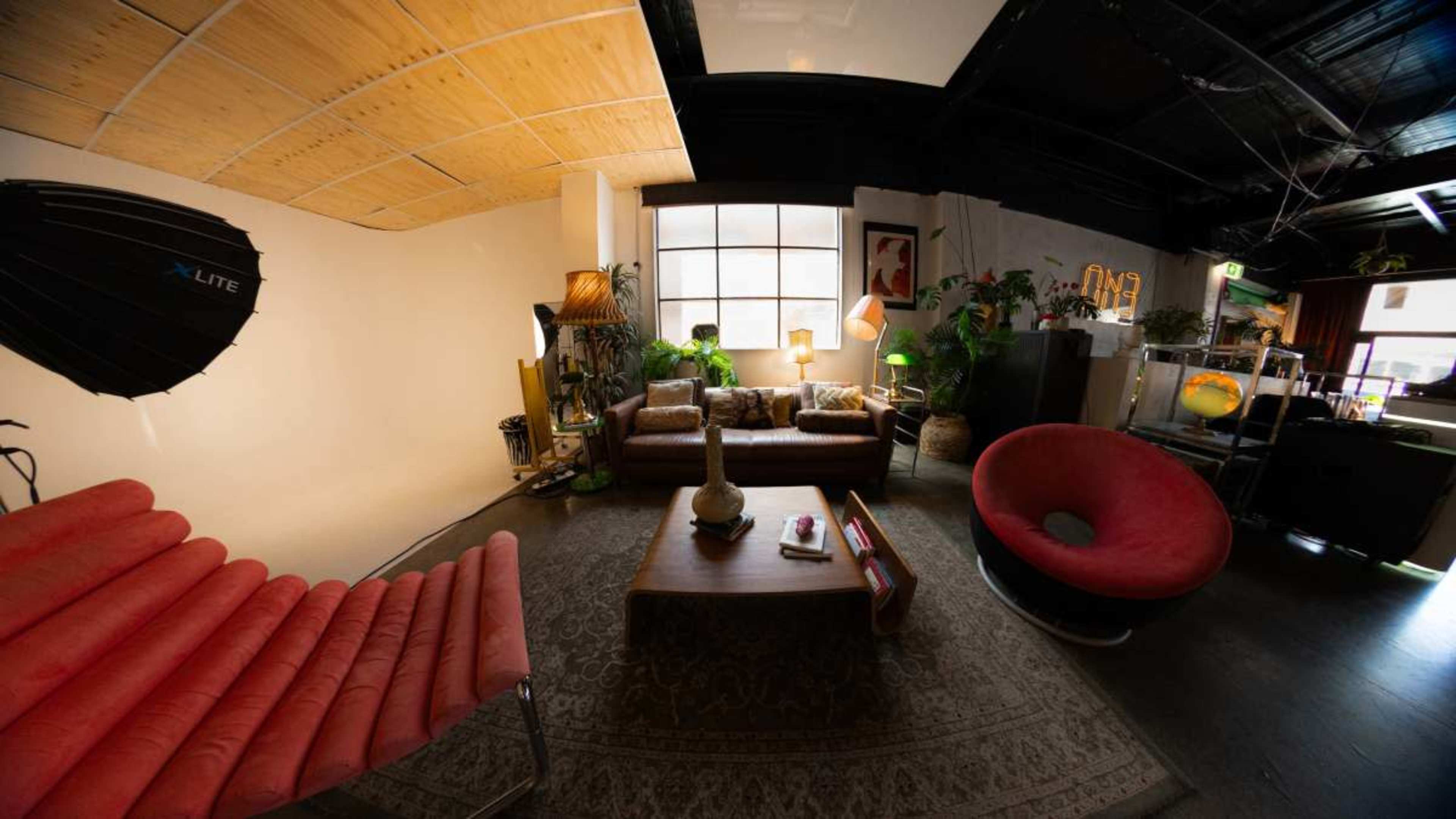 The image depicts a cozy lounge area featuring a pink sofa, brown seating, a coffee table, various plants, and a large window.