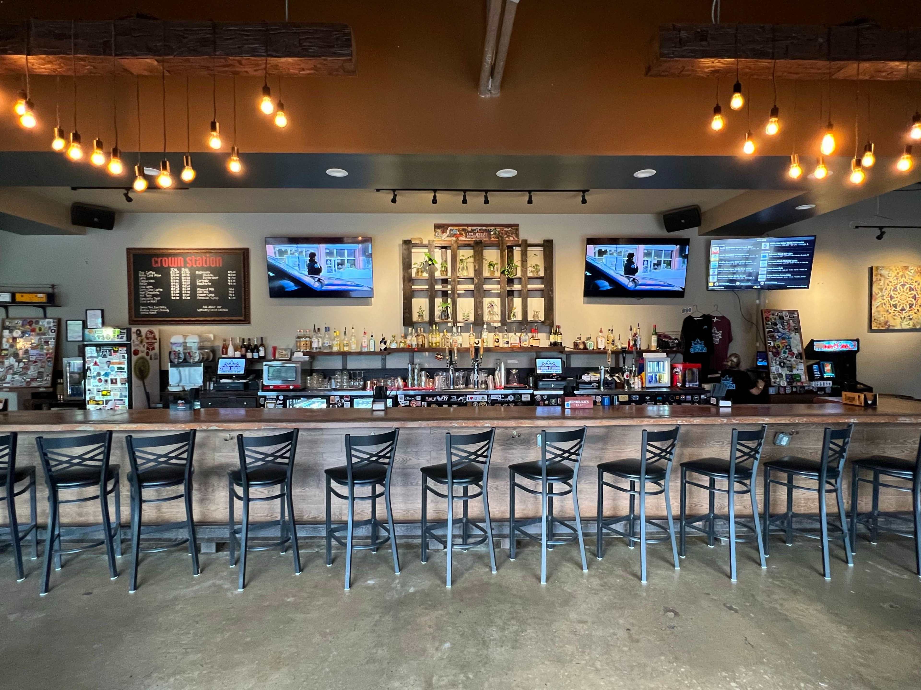 A well-lit bar area features a long wooden counter with high stools, shelves stocked with various liquor bottles, and multiple screens displaying menus and entertainment.