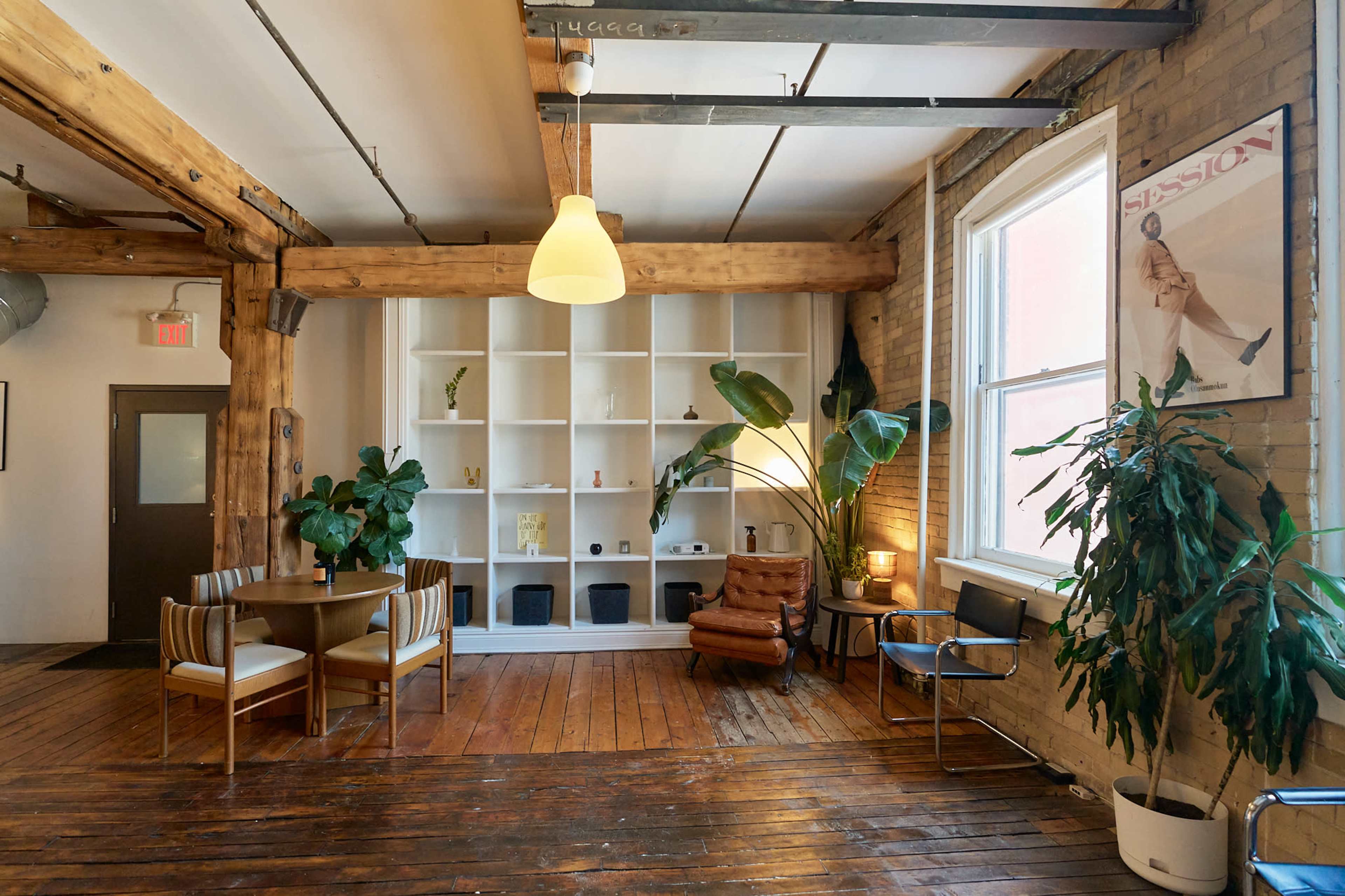 The image shows a spacious, warmly lit room featuring wooden beams, a circular dining table with chairs, tall indoor plants, and a white bookshelf against a brick wall.
