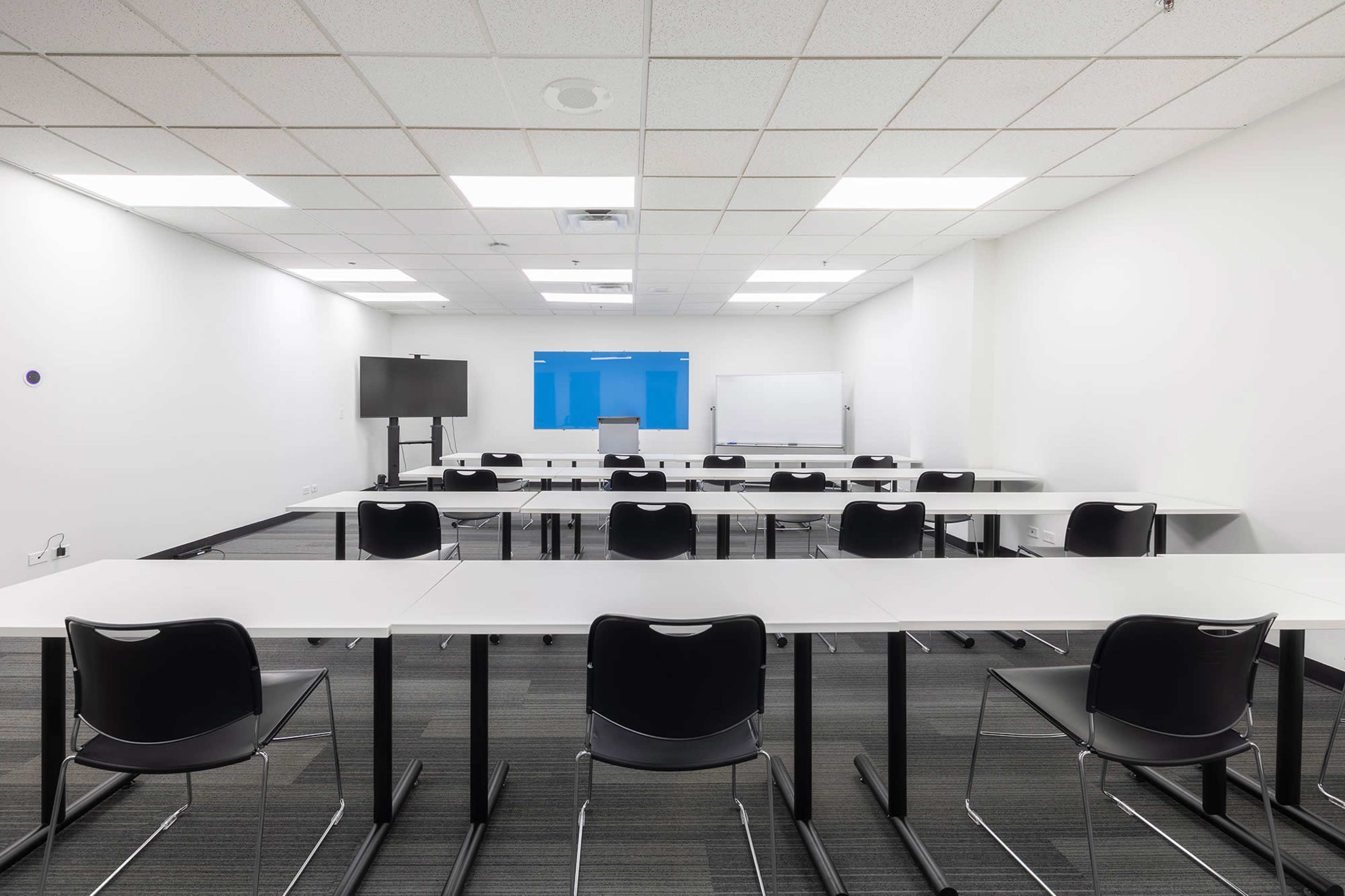 A modern classroom is set up with multiple tables and chairs, featuring a projector screen and whiteboard at the front.