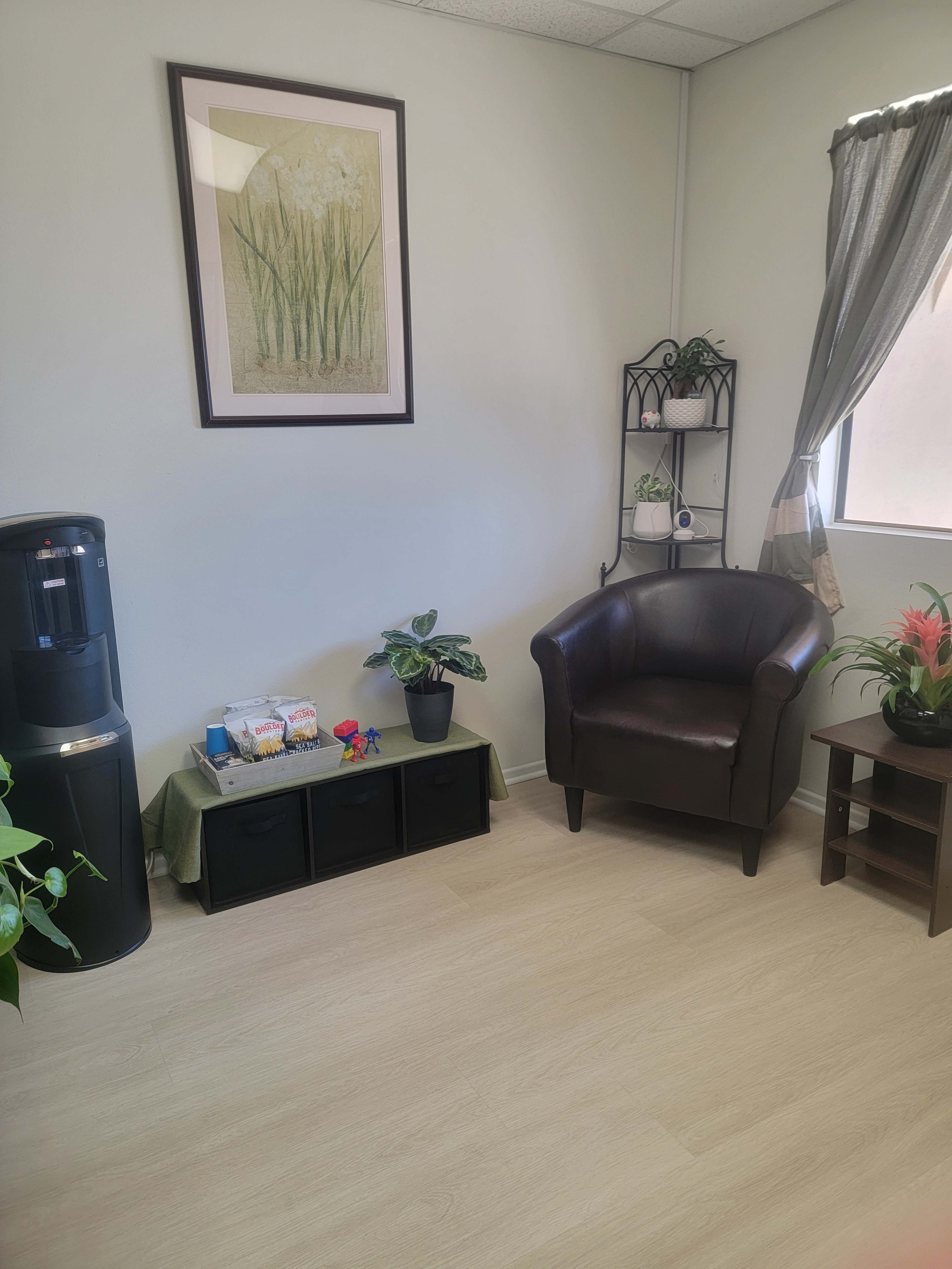 A small waiting room with a water cooler, a black armchair, a shelf with plants, and a framed botanical print on the wall.