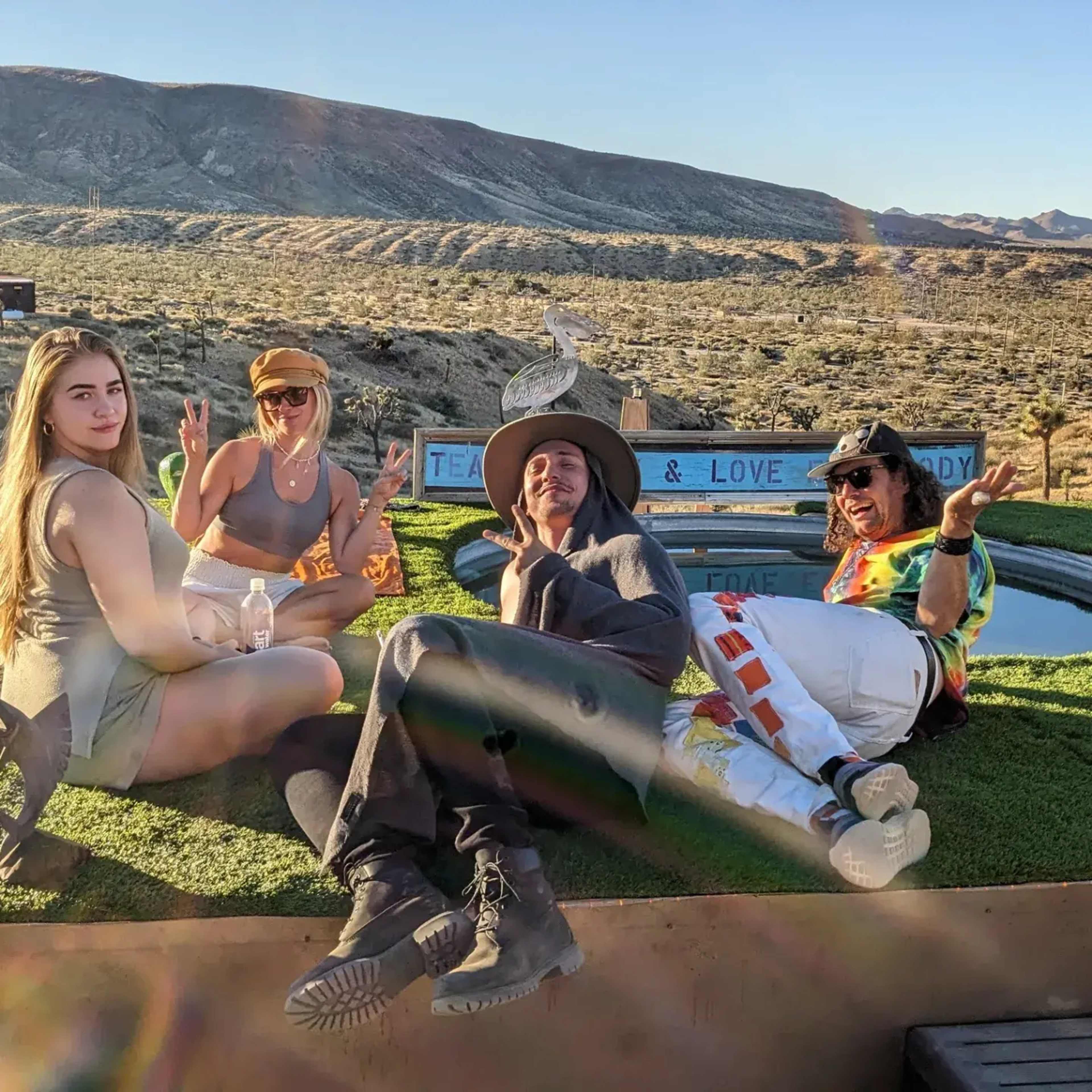 Four people relax outdoors near a swimming pool with a desert landscape in the background.