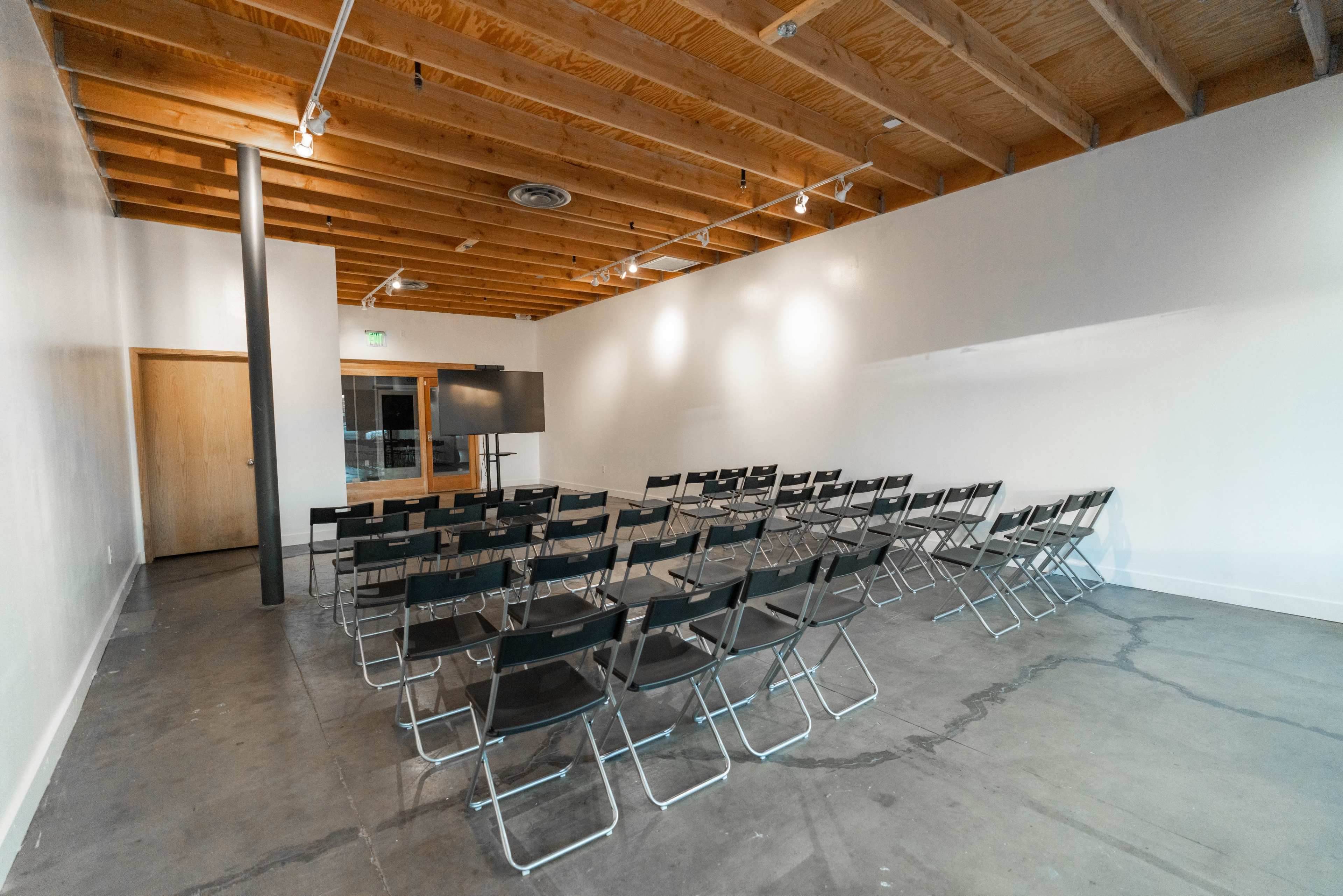 A row of black folding chairs is arranged facing a blank wall in a sparsely furnished meeting room with wooden beams.