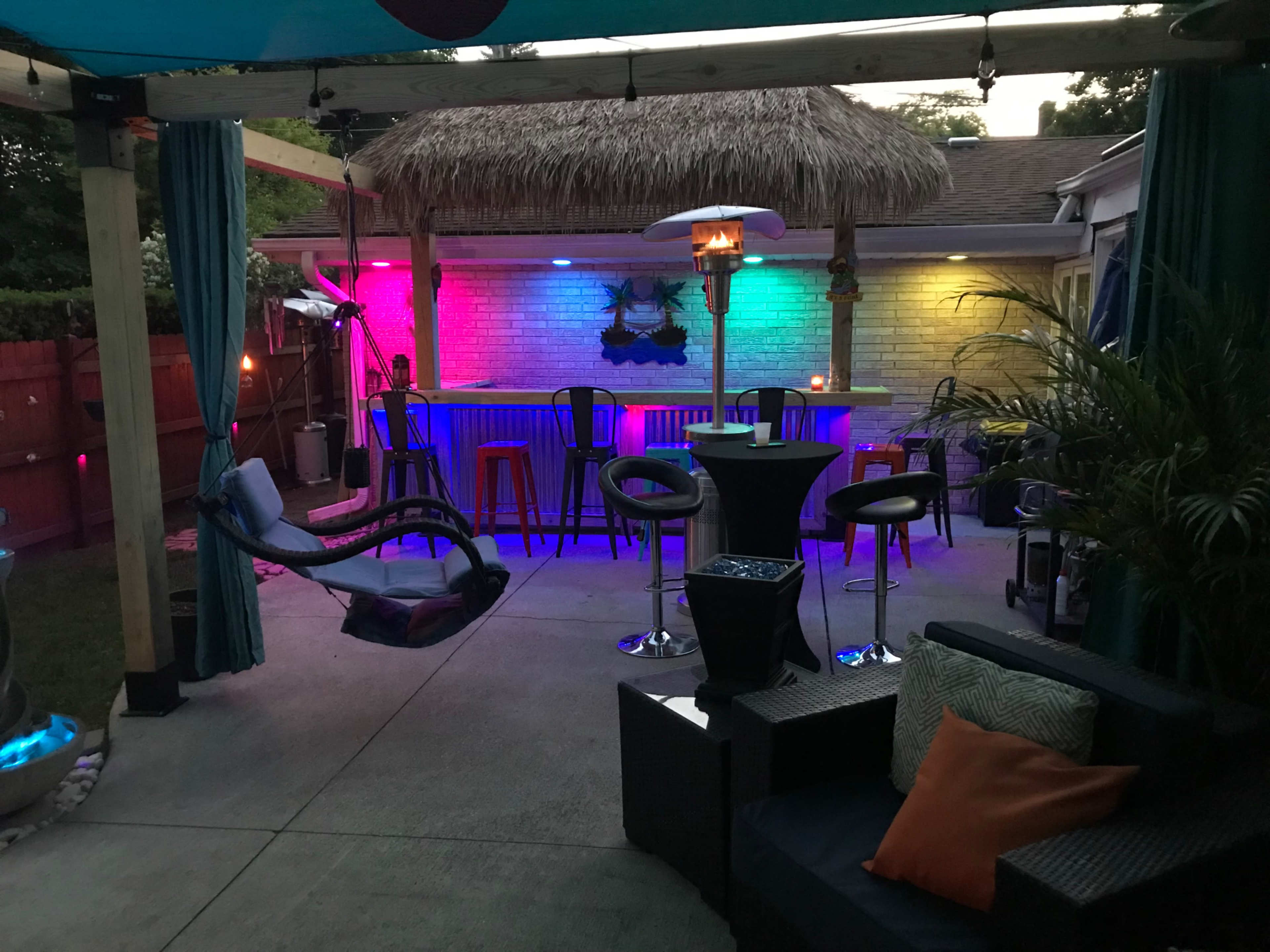 A backyard patio features a thatched-roof bar with colorful LED lighting, surrounded by seating areas and a hammock.