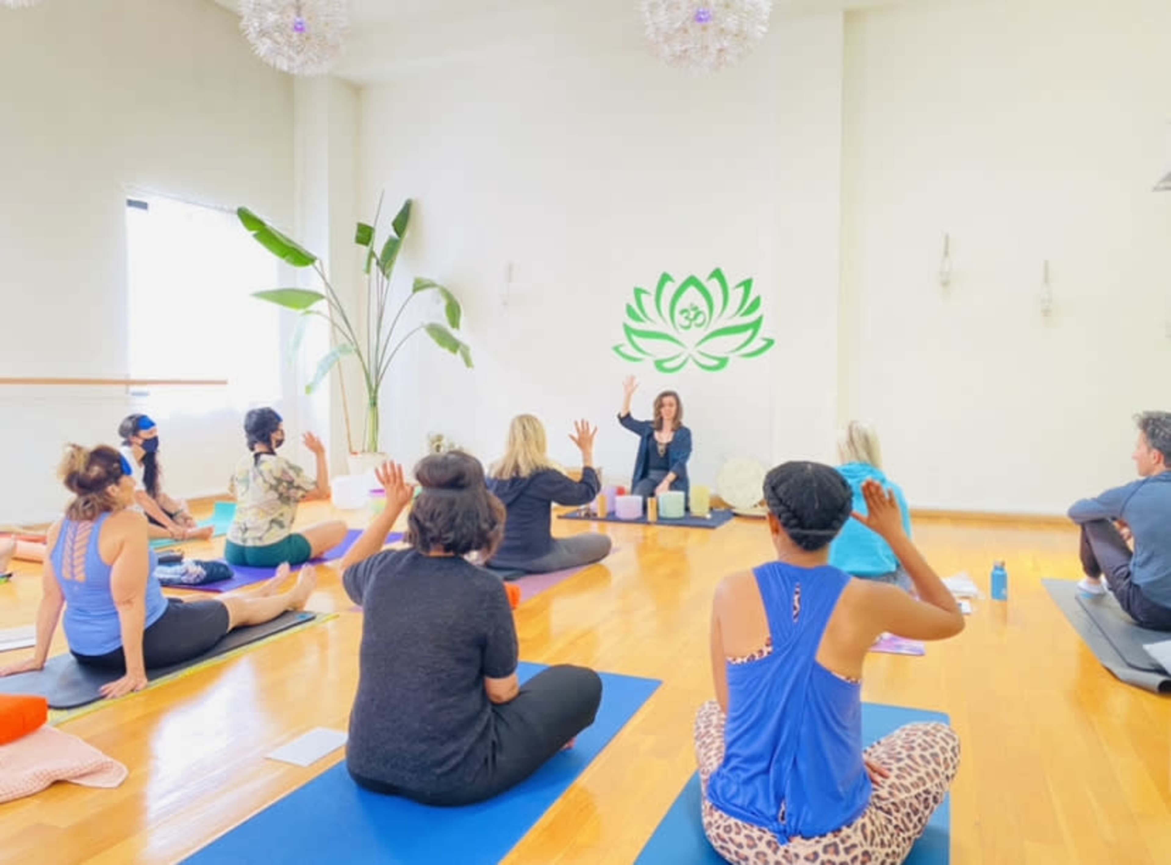 A group of people sits on yoga mats in a bright studio, attentively facing an instructor who is leading a class.