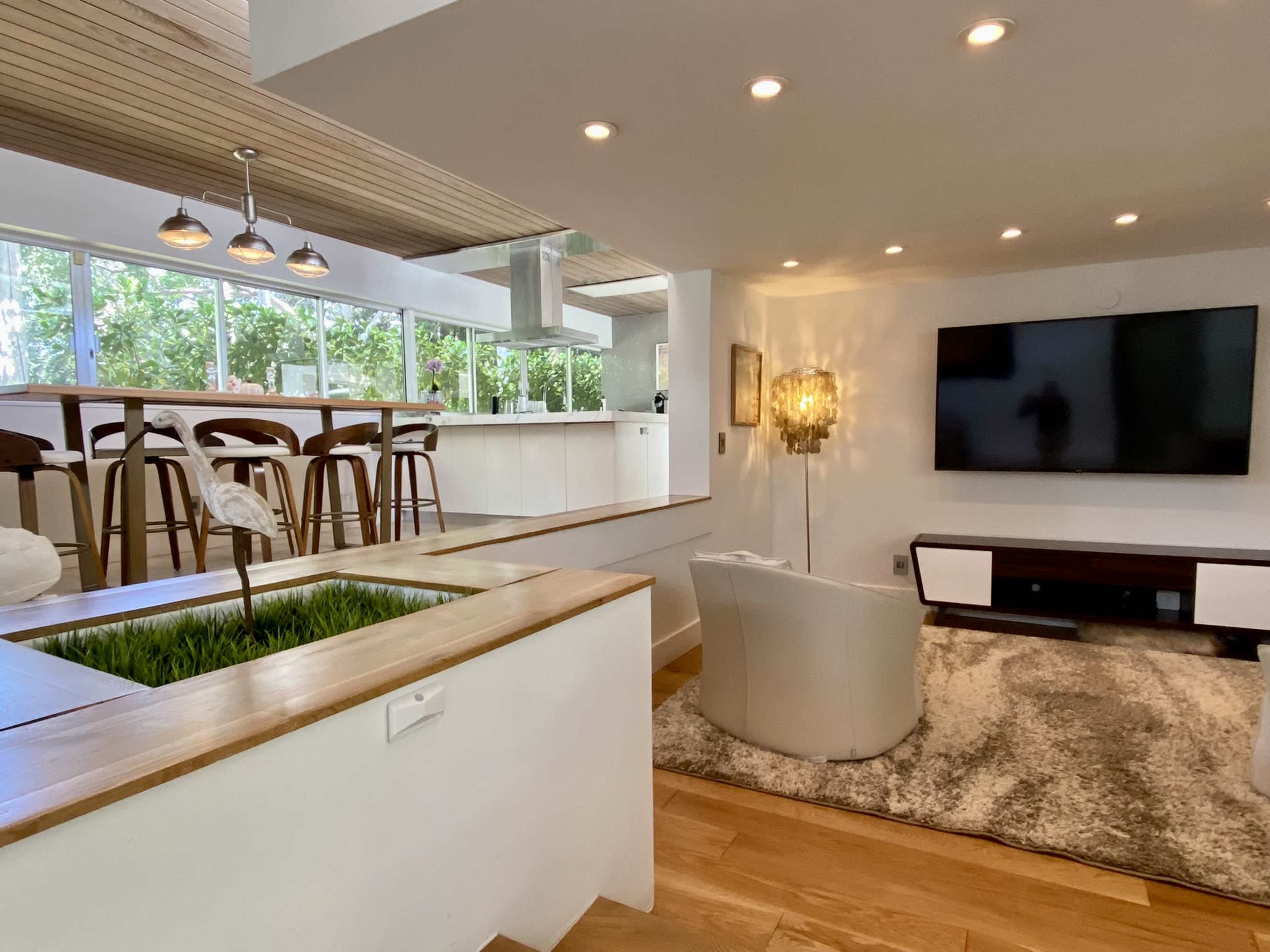 The image shows a modern living room featuring a white armchair, a large television mounted on the wall, and a kitchen bar area with three stools, all illuminated by ample natural light from large windows.