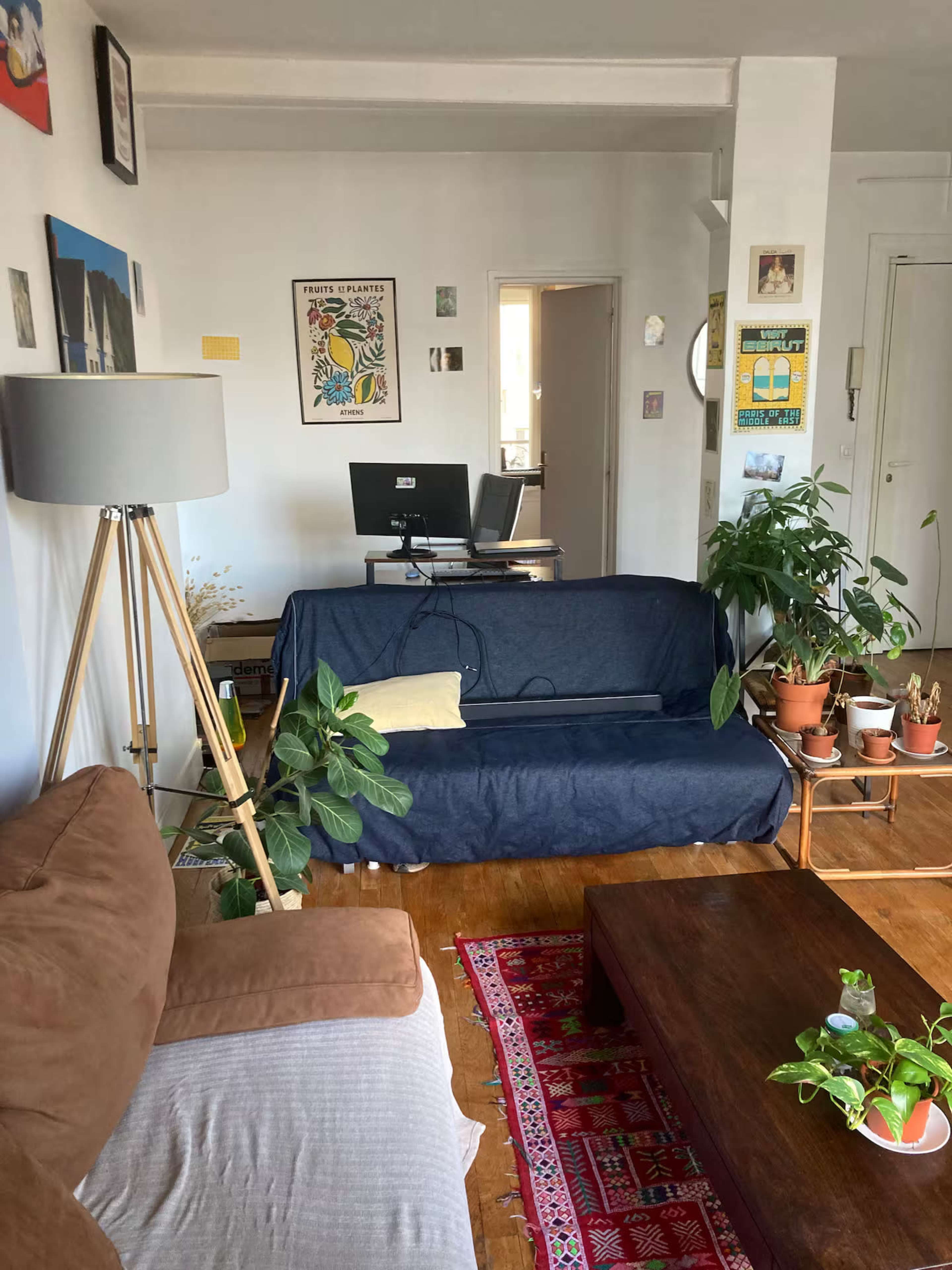 The image shows a cozy living room with a brown couch, a blue-covered sofa, a wooden coffee table, a floor lamp, and various plants.
