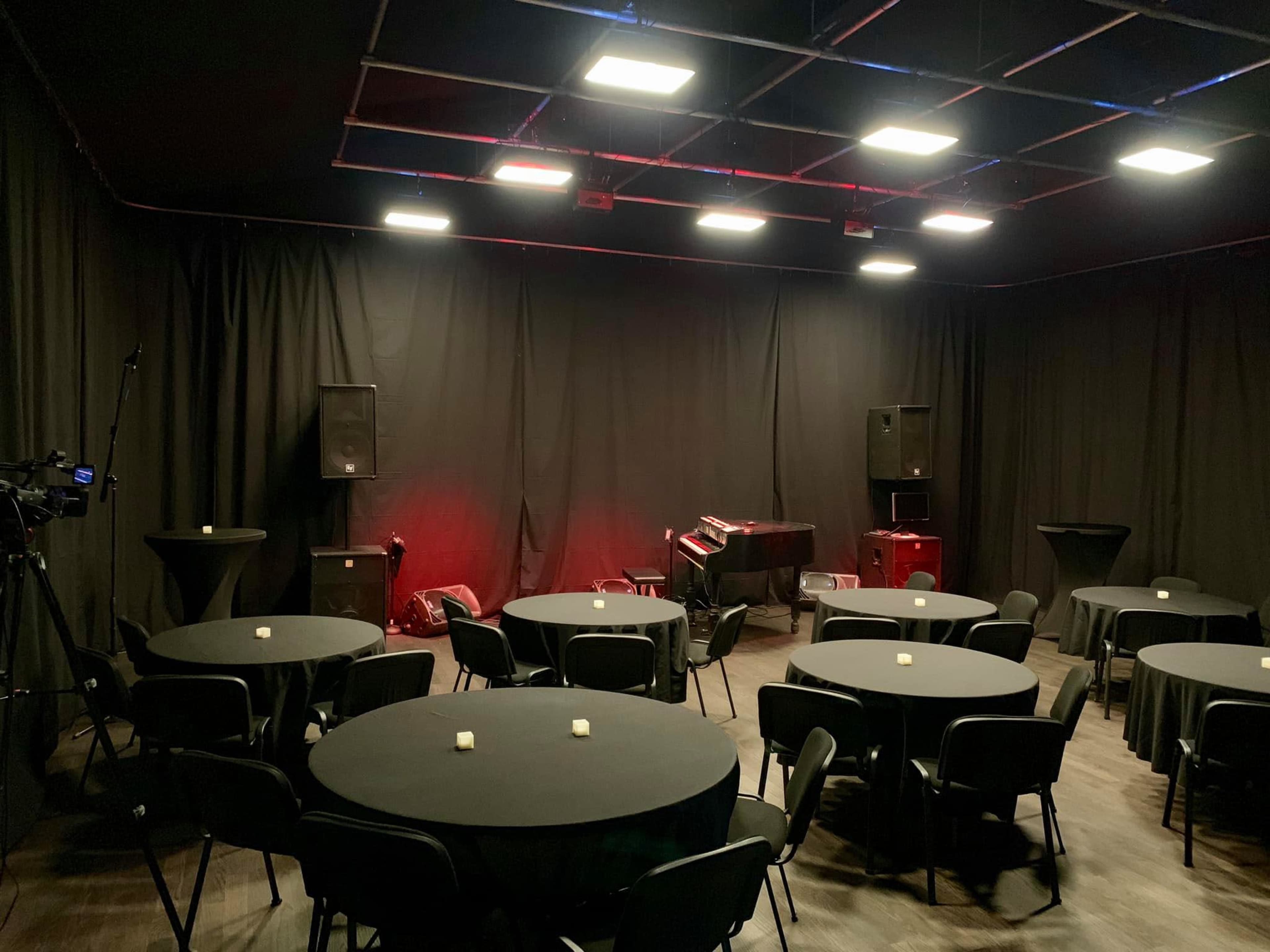 A black-walled performance space is set up with round tables and a grand piano, flanked by speakers and a camera.