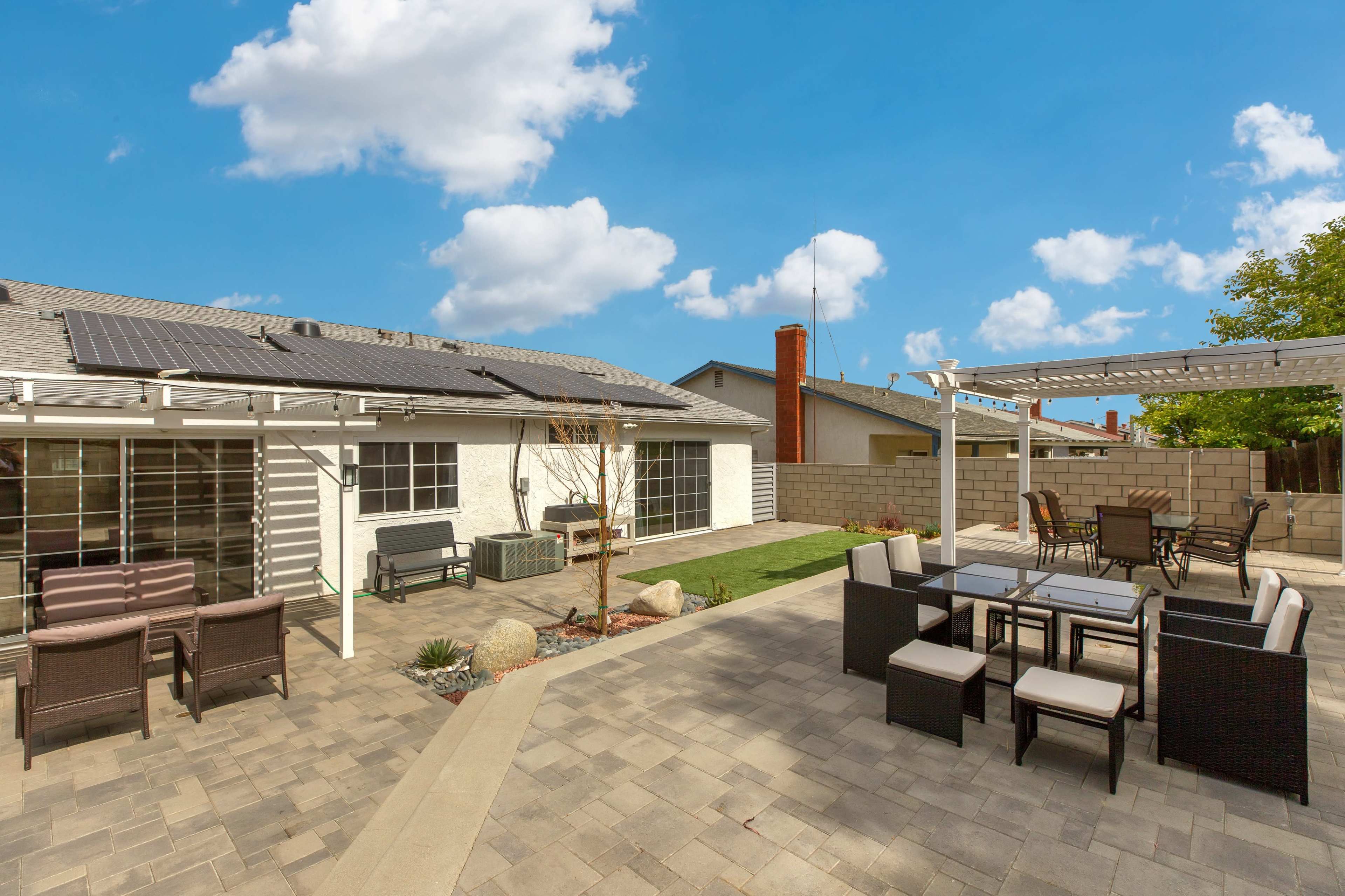 The image shows a backyard with a paved area featuring outdoor furniture, a grassy section, and solar panels installed on the roof of a house.