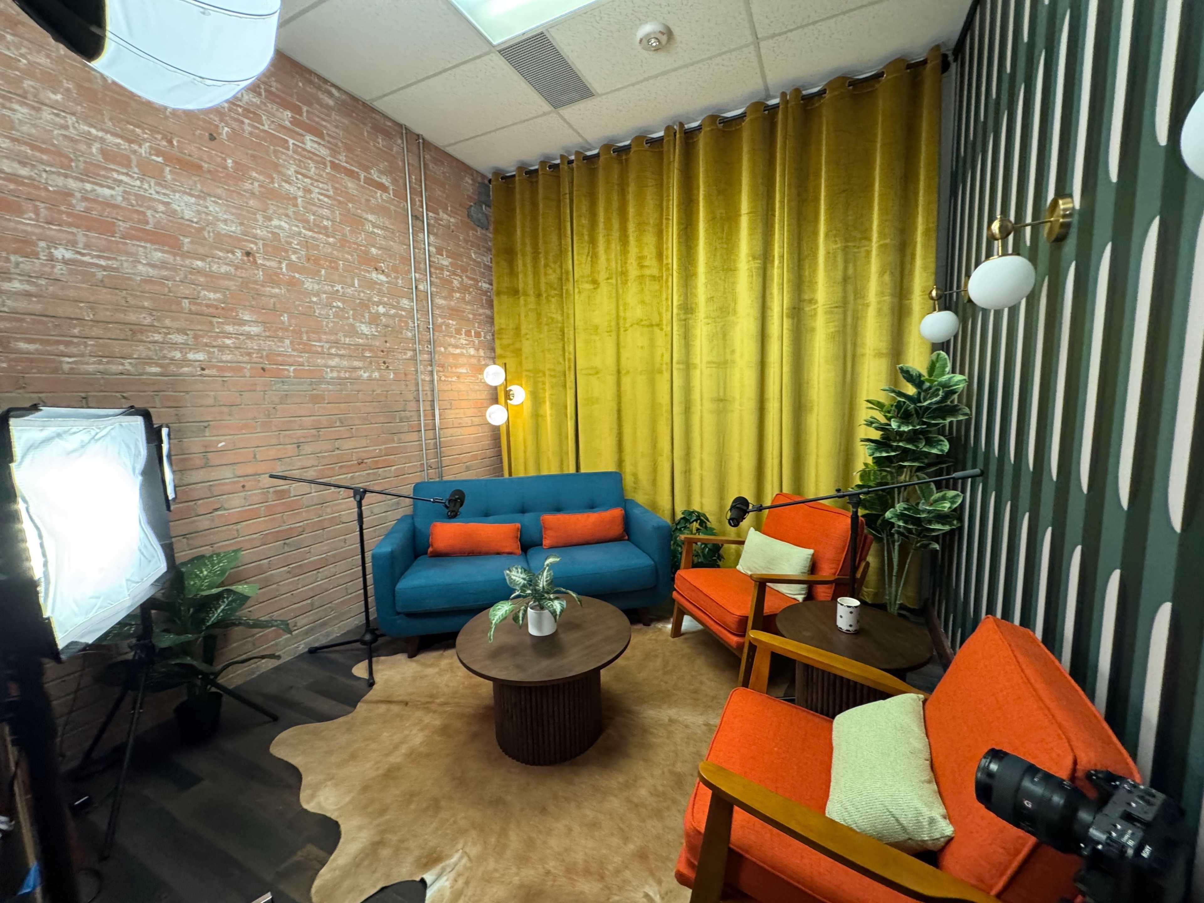 The image shows a cozy indoor setting with a teal sofa, two orange chairs, a round table, and a yellow curtain against a brick wall.
