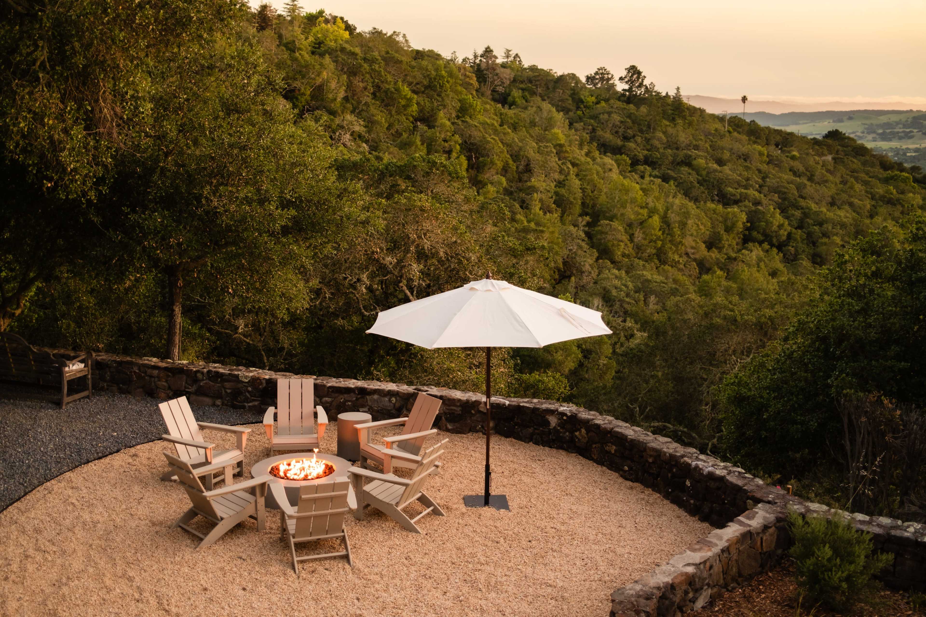 A circular area with several white Adirondack chairs encircles a fire pit, situated on a gravel surface with a large umbrella, overlooking a wooded hillside during sunset.