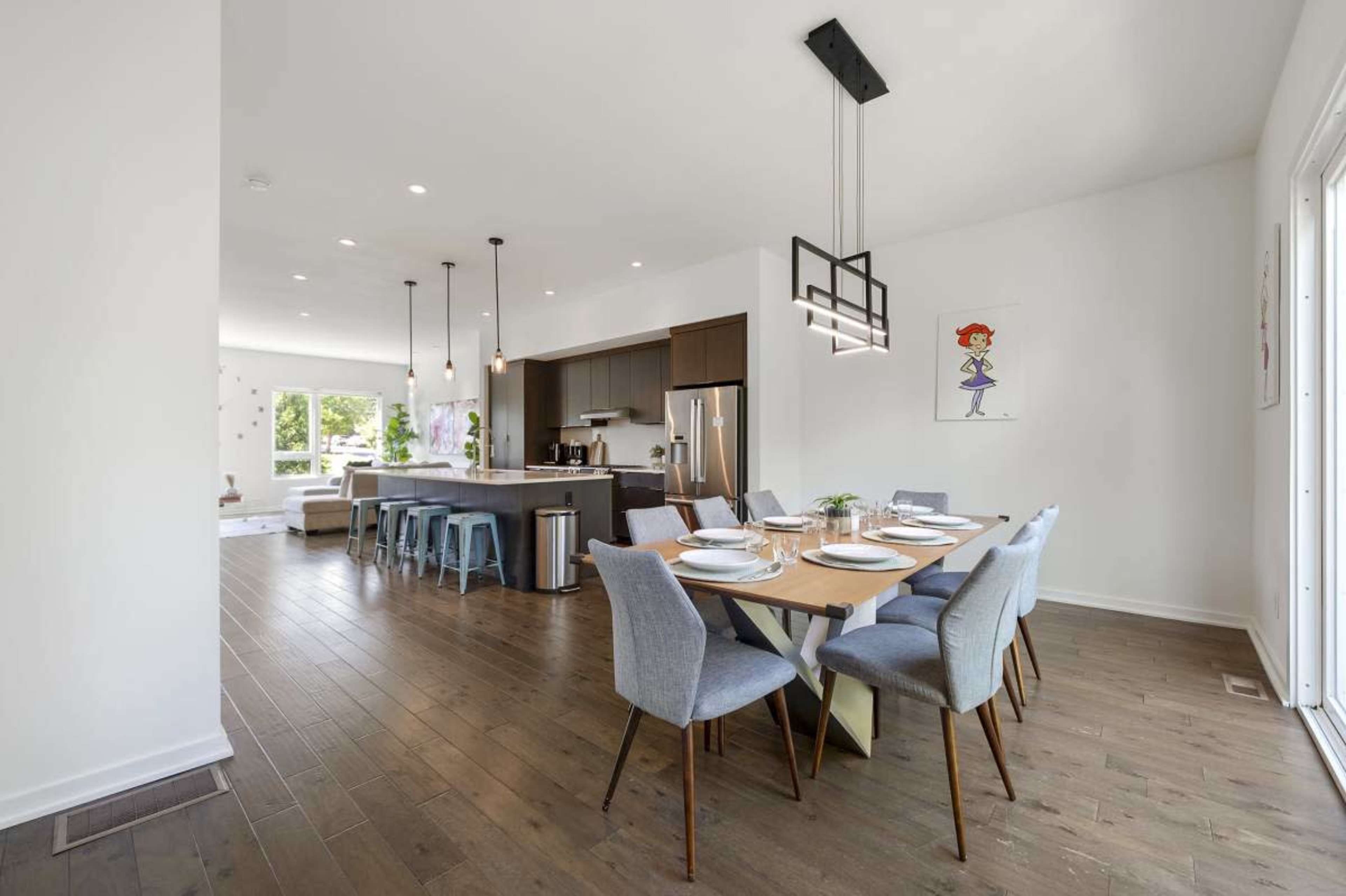 The image shows a modern kitchen and dining area with wooden flooring, a large table set for a meal, and sleek cabinets.