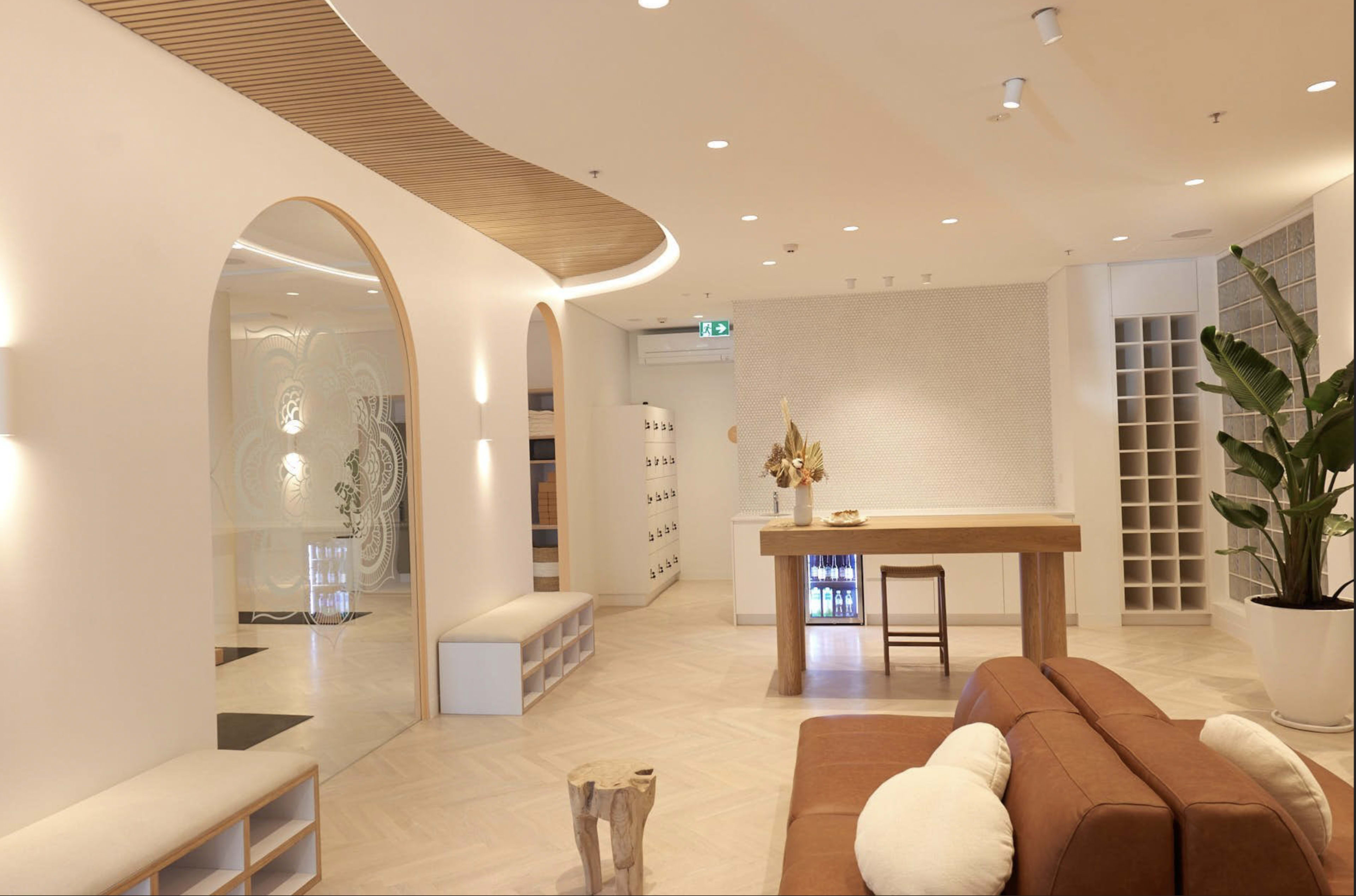 A modern interior space featuring a beige couch, wooden furniture, greenery, and light-colored walls with curved architectural details.
