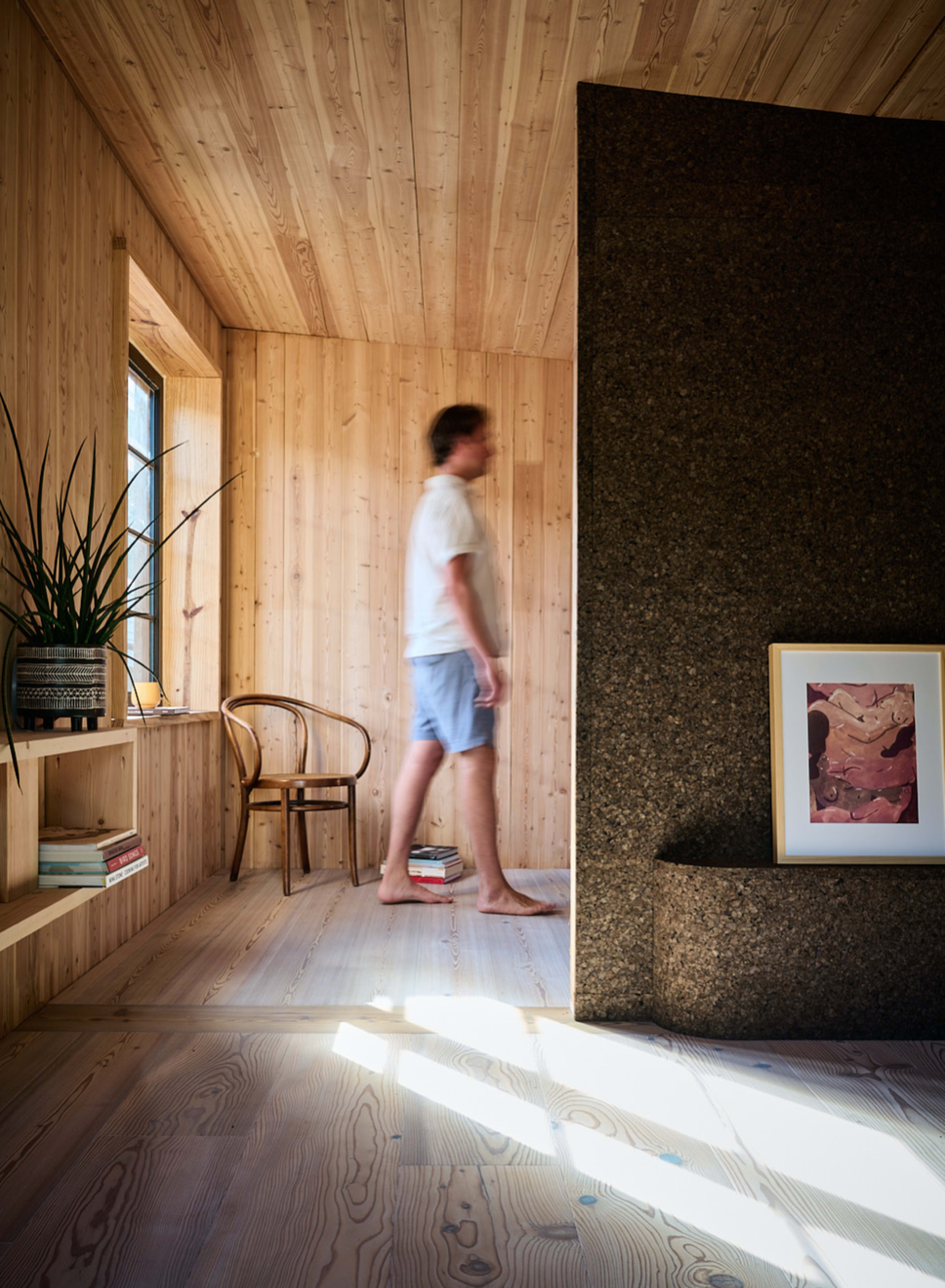 A person walks through a wooden interior space featuring a large stone wall, shelves, and a framed artwork.