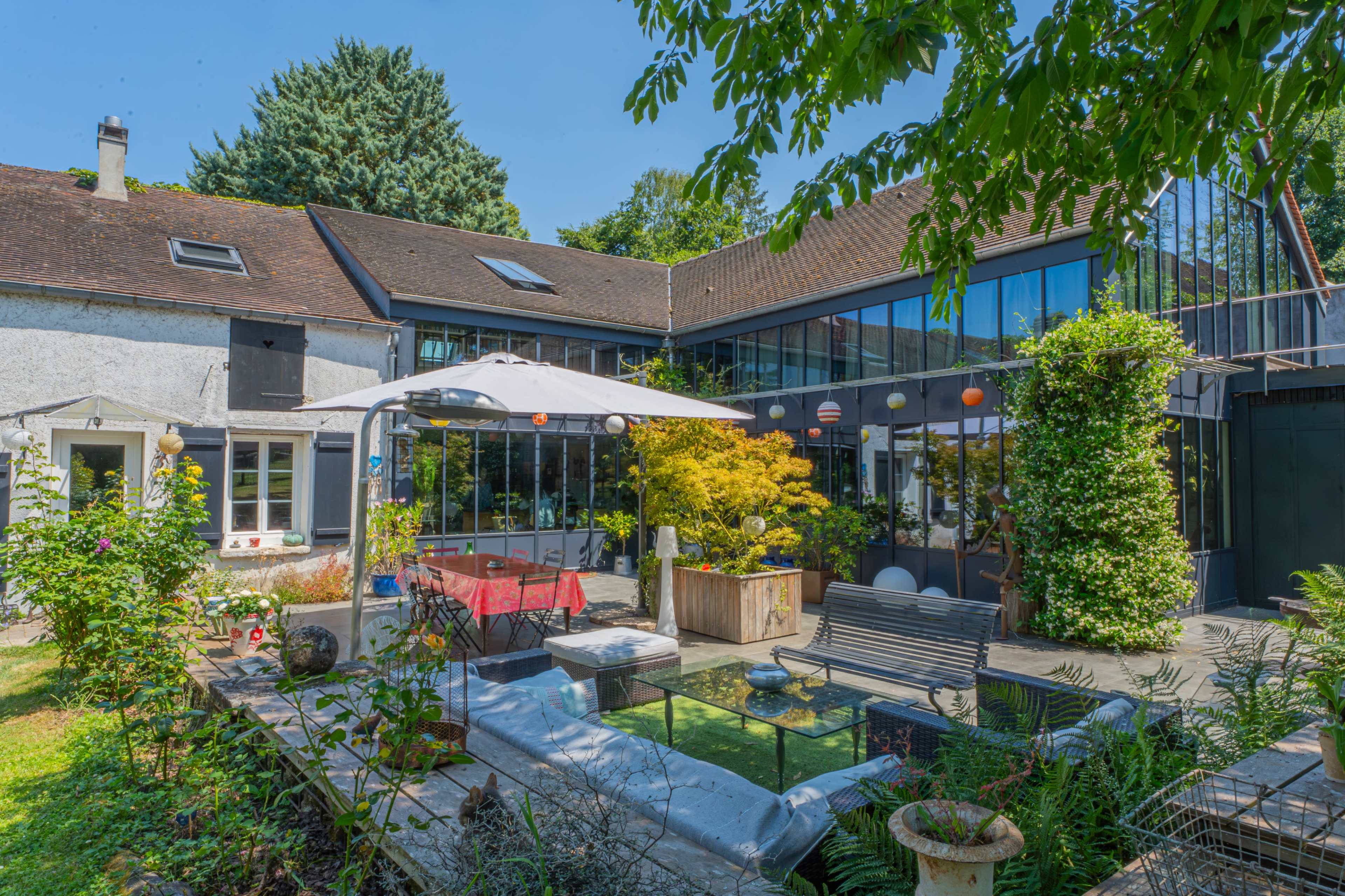 The image shows a serene backyard with a large patio area surrounded by greenery, featuring a mix of seating arrangements and a shaded dining space.
