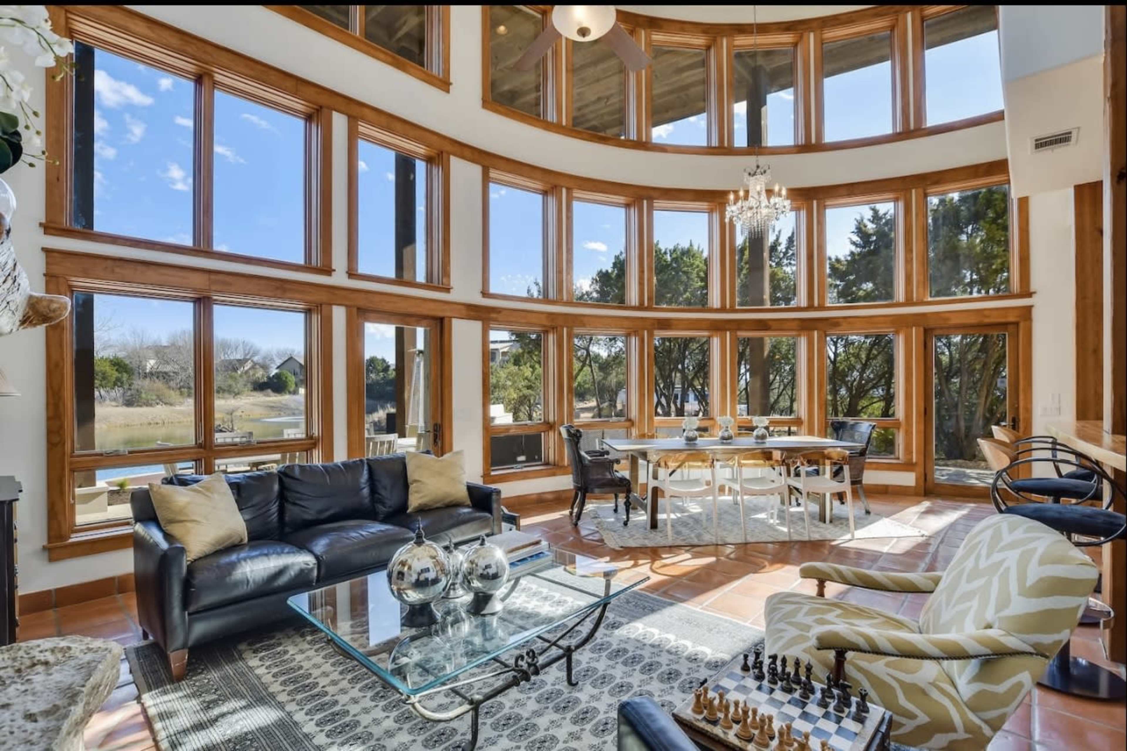 The image shows a spacious living room with large windows allowing natural light, featuring a black couch, a glass coffee table, and a dining area with a round table.