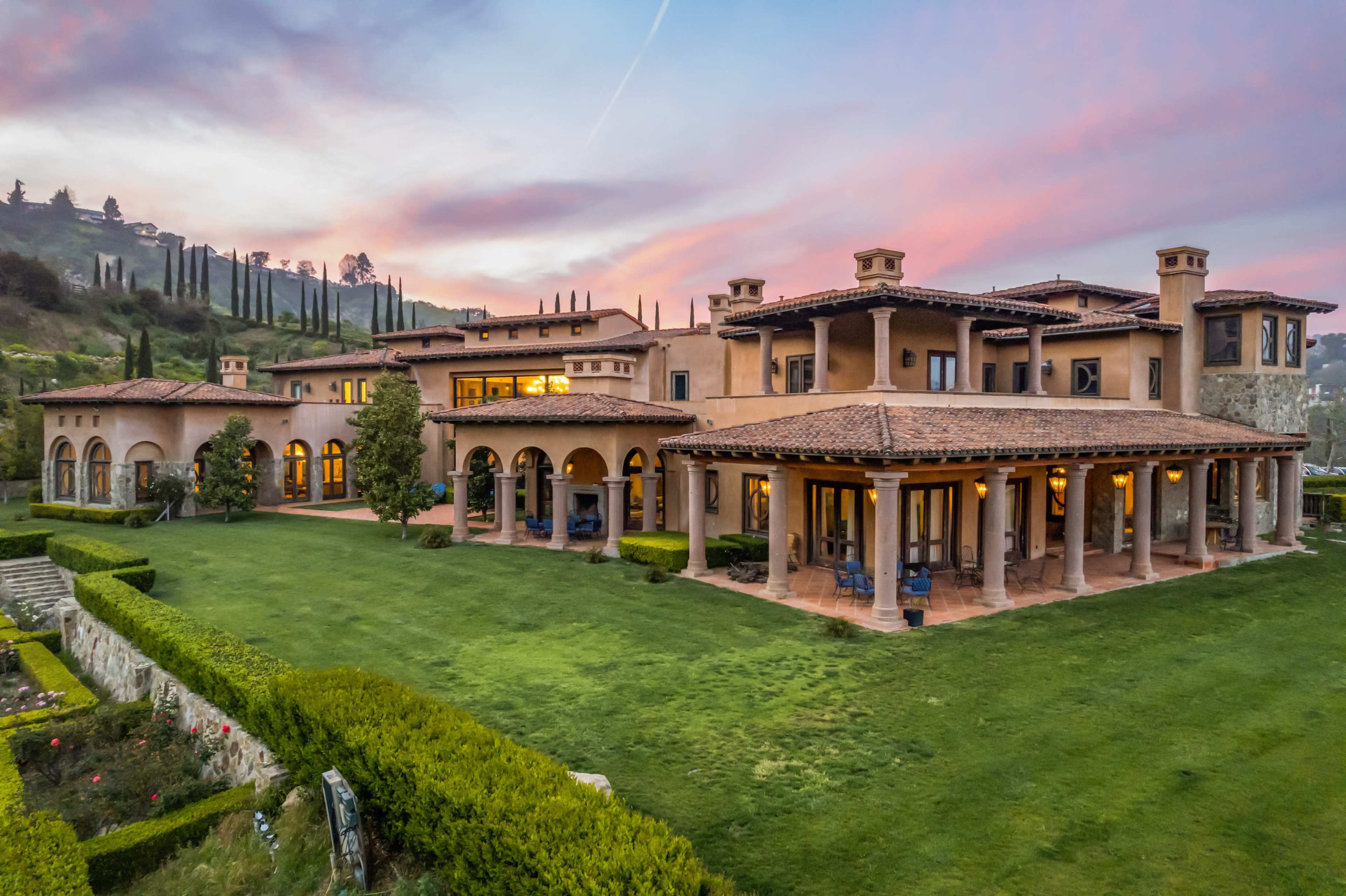 A large, sprawling Italian-style villa is set against a colorful sunset, surrounded by manicured lawns and gardens.
