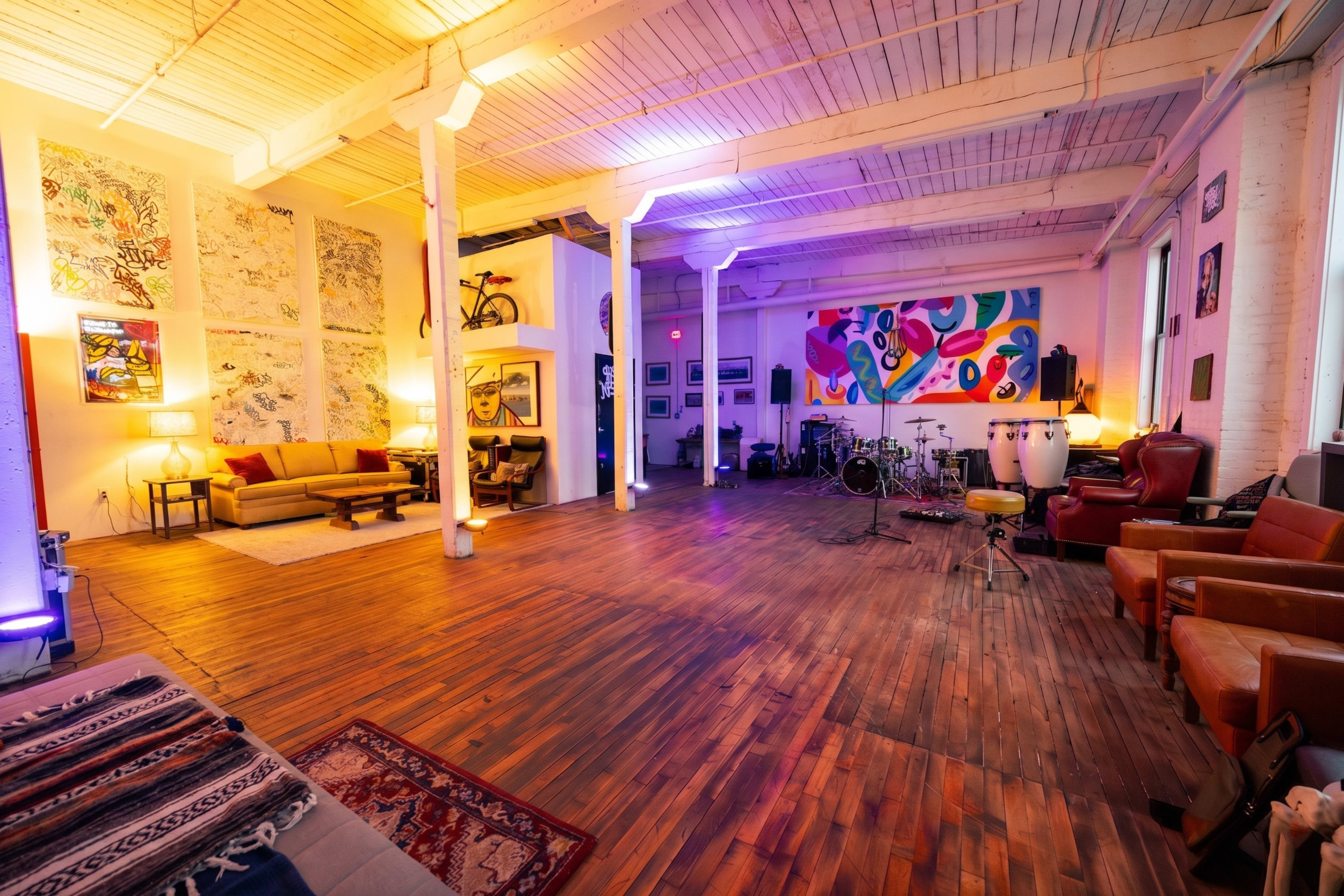 The image shows a spacious, well-lit room with wooden floors, featuring a seating area with couches, vibrant artwork on the walls, and various musical instruments arranged in one corner.