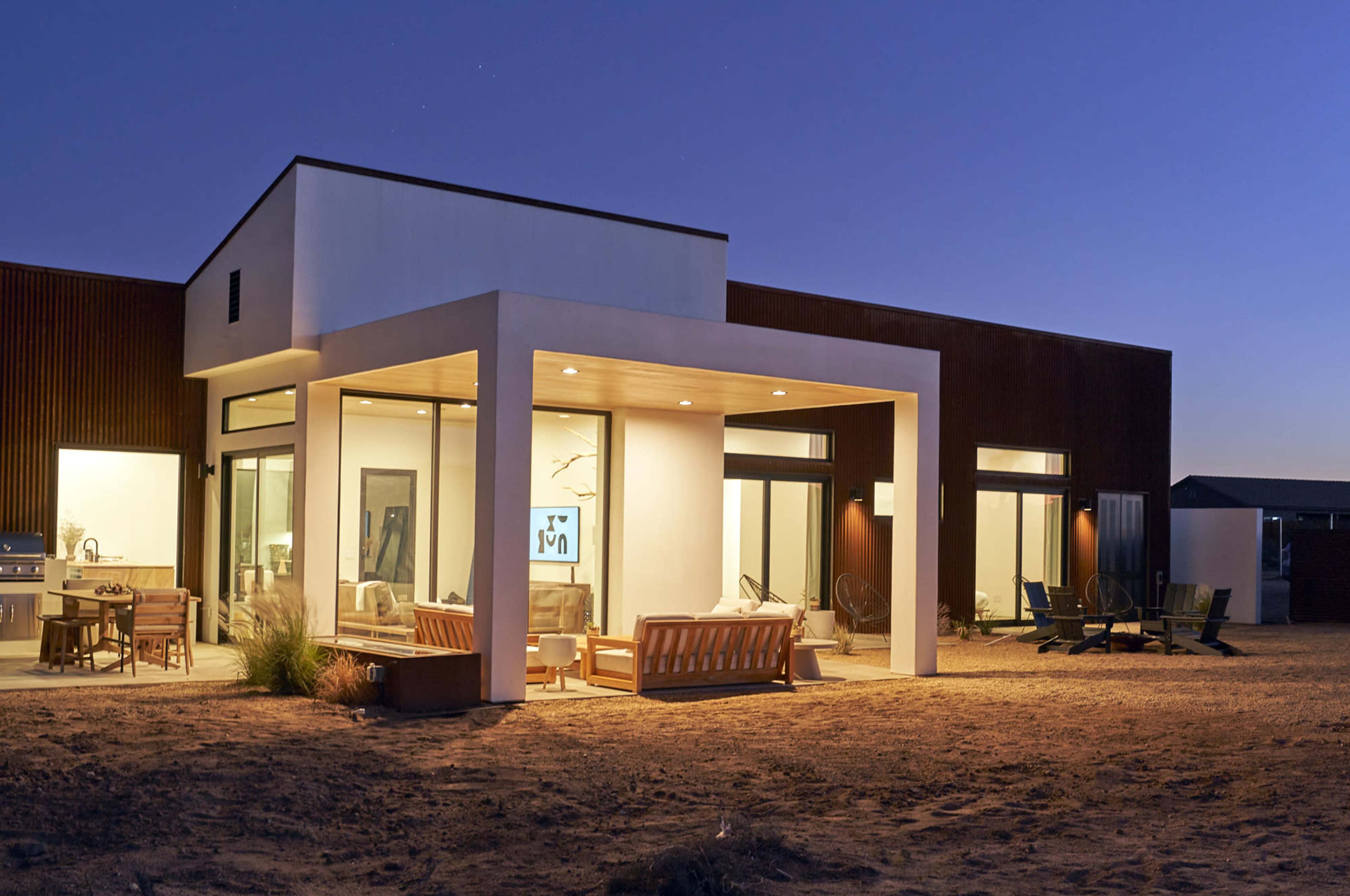 The image shows a modern single-story house with a large covered patio overlooking a sandy landscape at dusk.