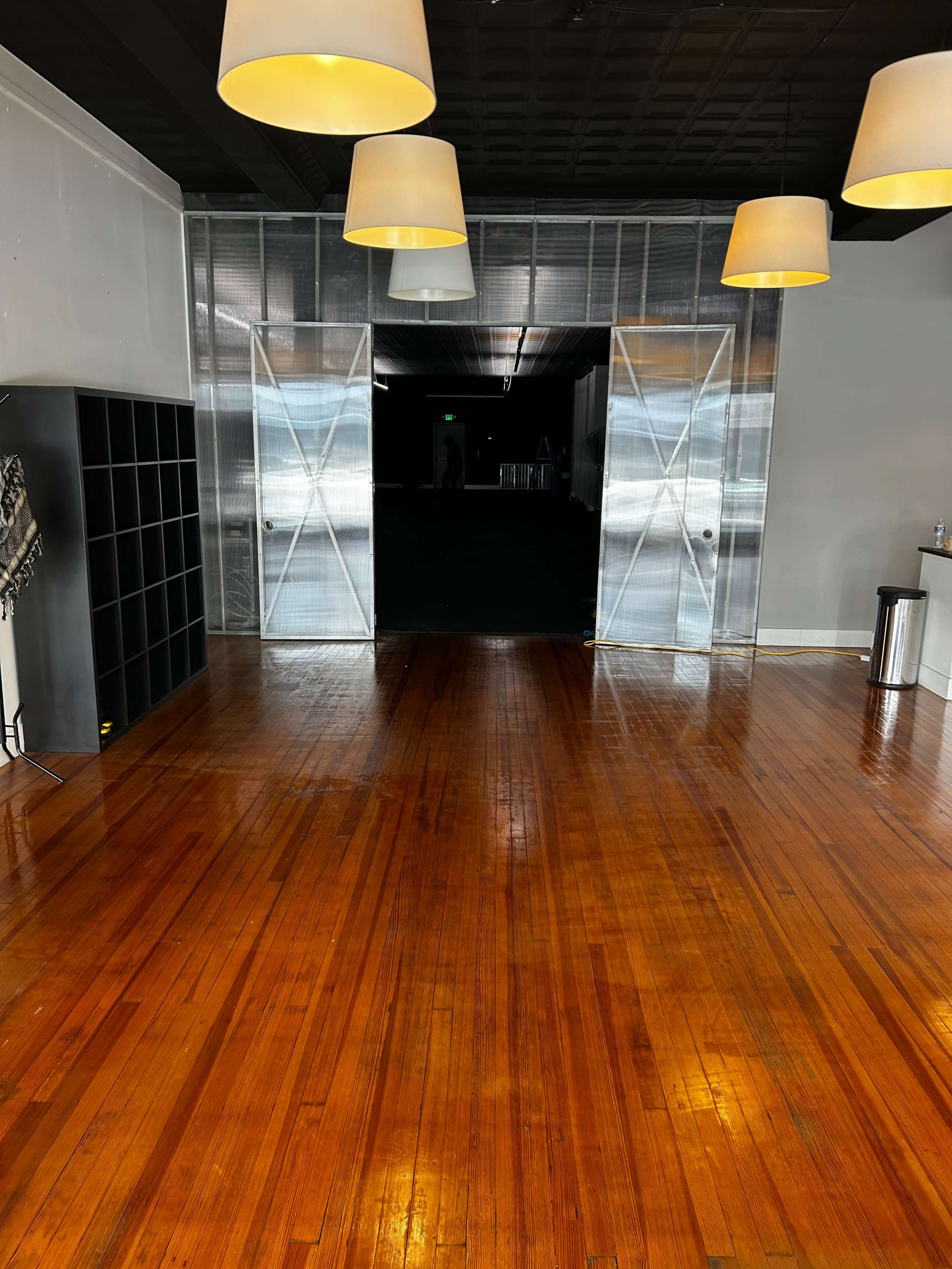 The image shows a spacious room with polished wooden floors, illuminated by hanging lamps, leading to large open doors at the far end.