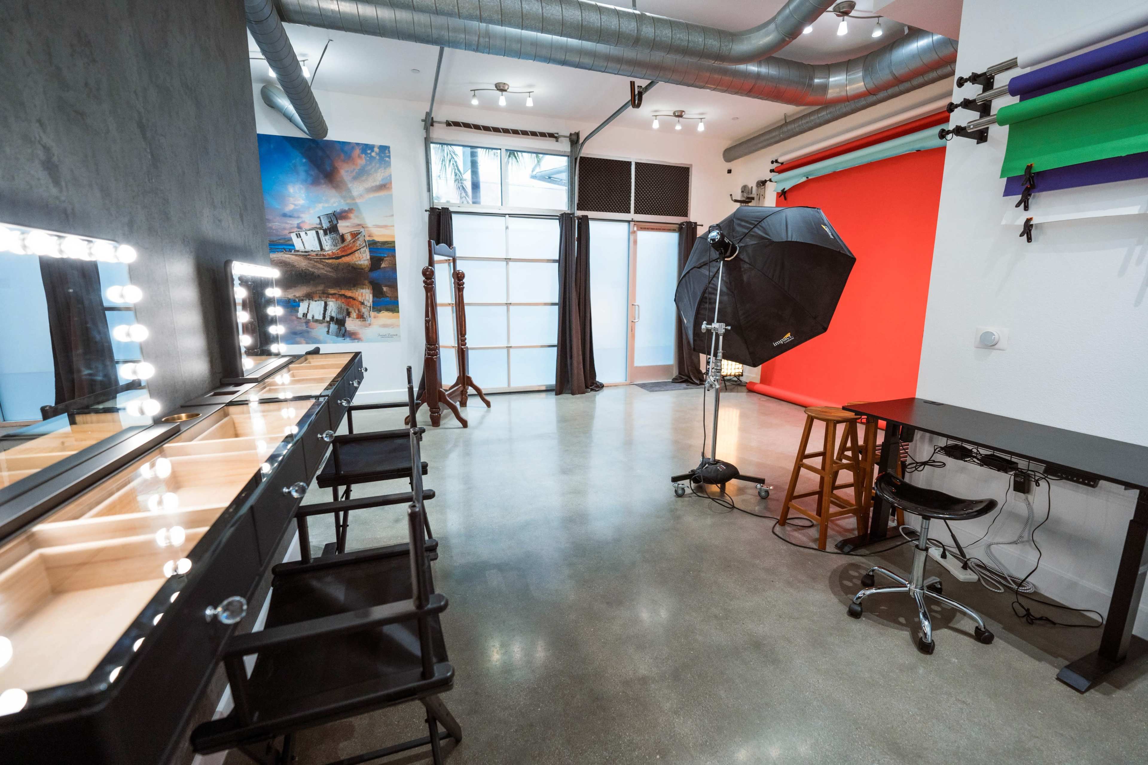 A photography studio featuring a makeup station with illuminated mirrors, a backdrop setup with colored rolls, and a softbox lighting device.
