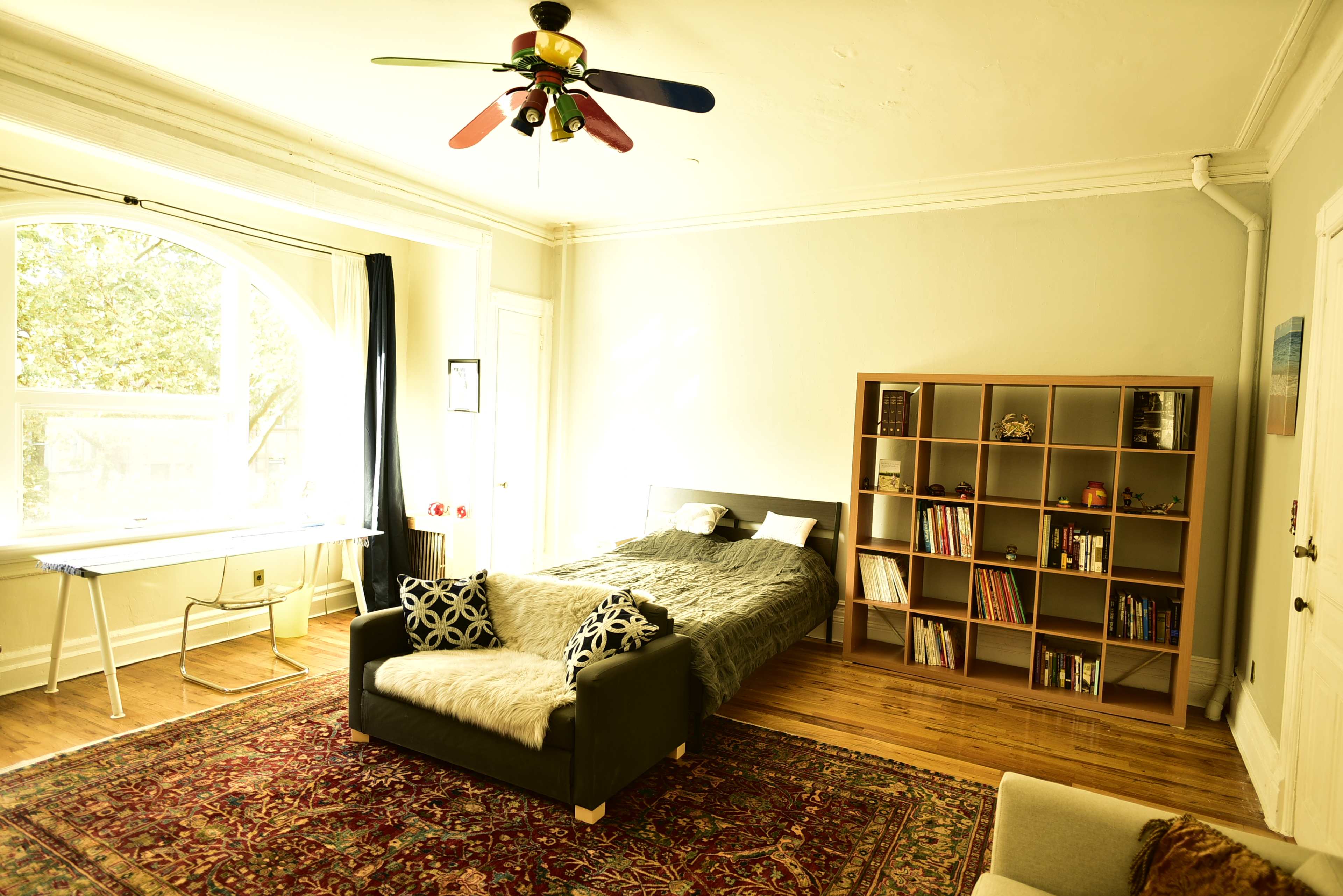 The room features a bed, a bookshelf with various items, a desk by a large window, and a couch on a patterned rug.