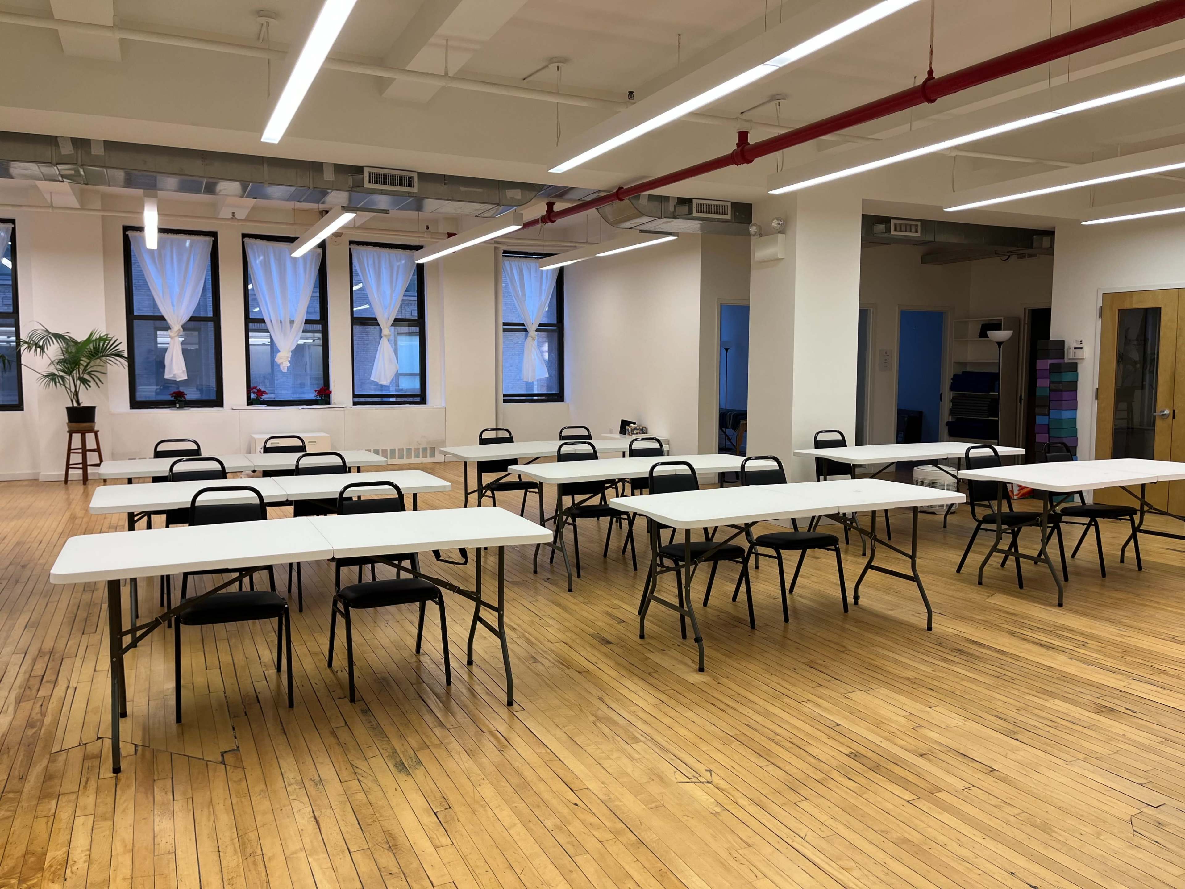 A spacious room with wooden floors, featuring several rows of folding tables and chairs, and large windows with sheer curtains.