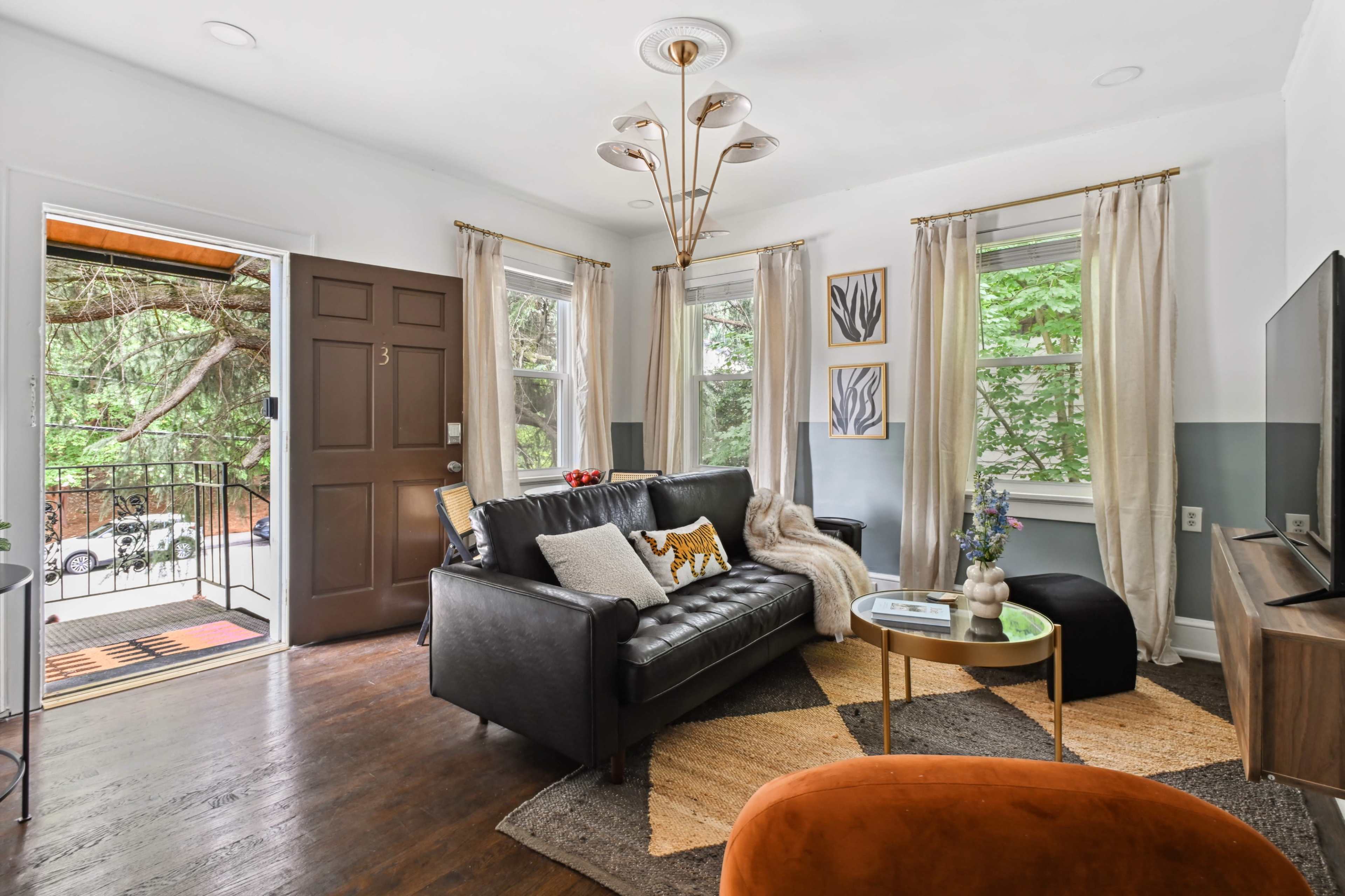 The living room features a black leather sofa, a round coffee table, and large windows that overlook a tree-filled balcony.
