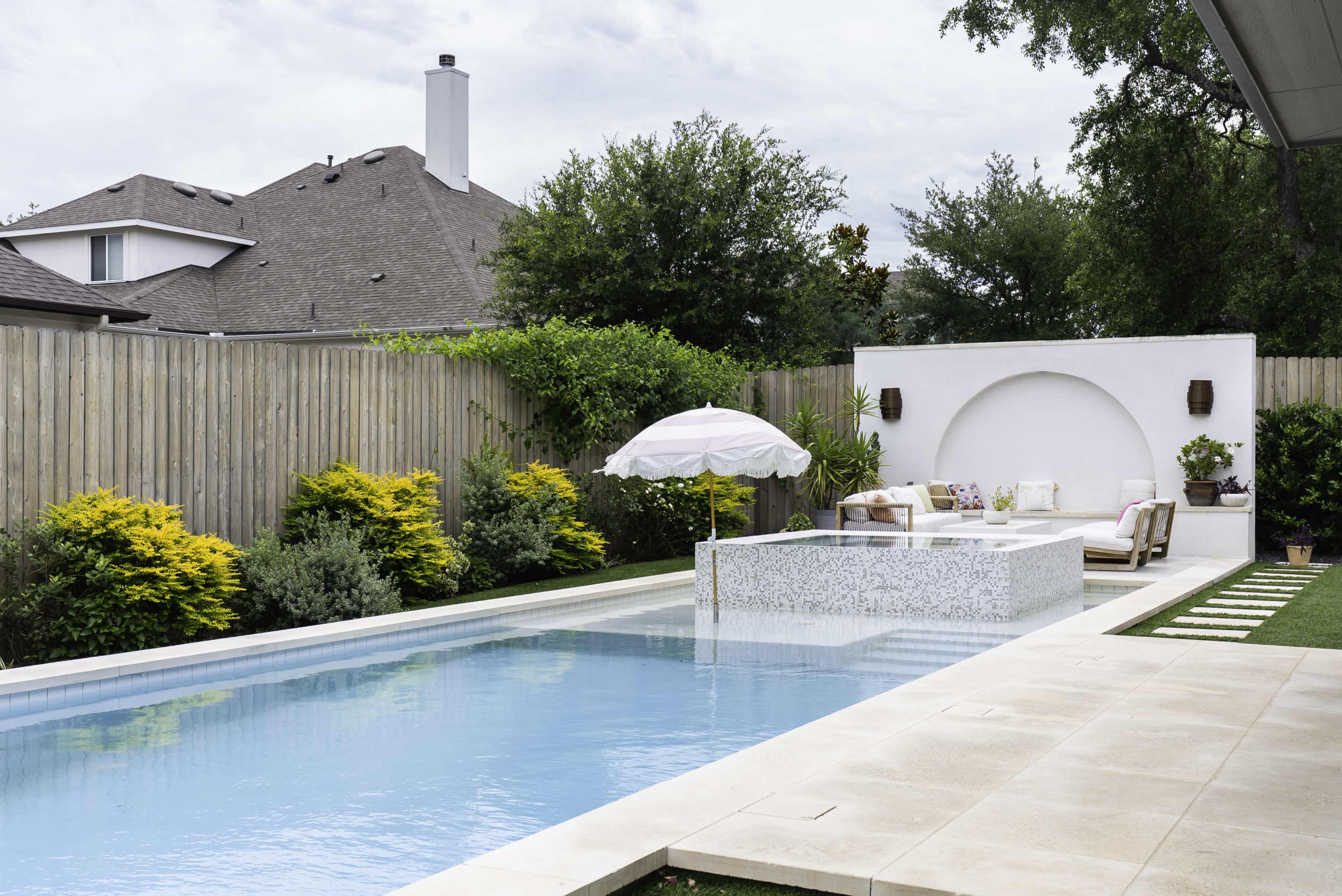 A rectangular swimming pool is surrounded by landscaped greenery and features a small seating area with an umbrella beside it.