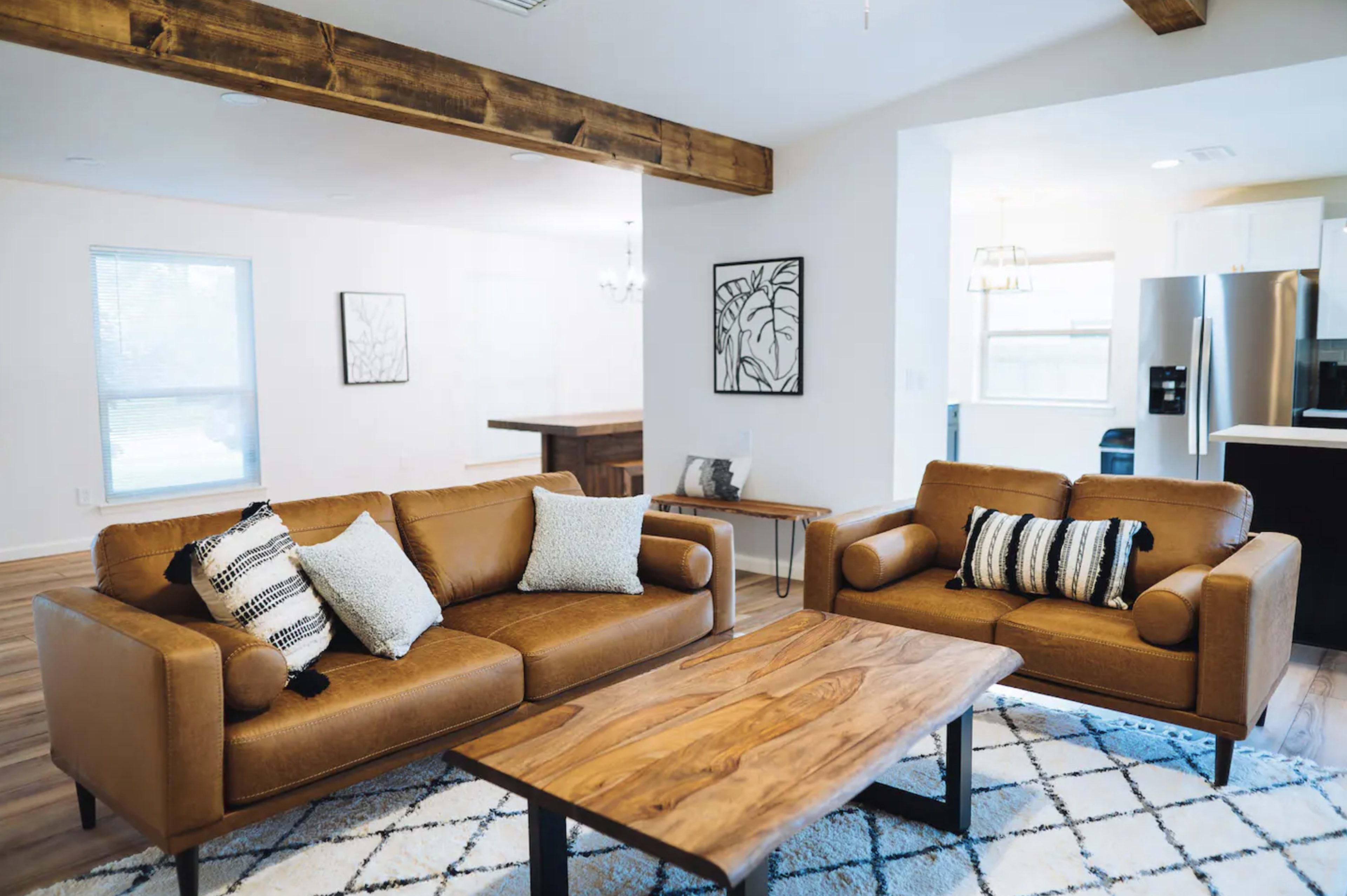A modern living room features brown leather sofas with decorative pillows, a wooden coffee table, and a dining area in the background.