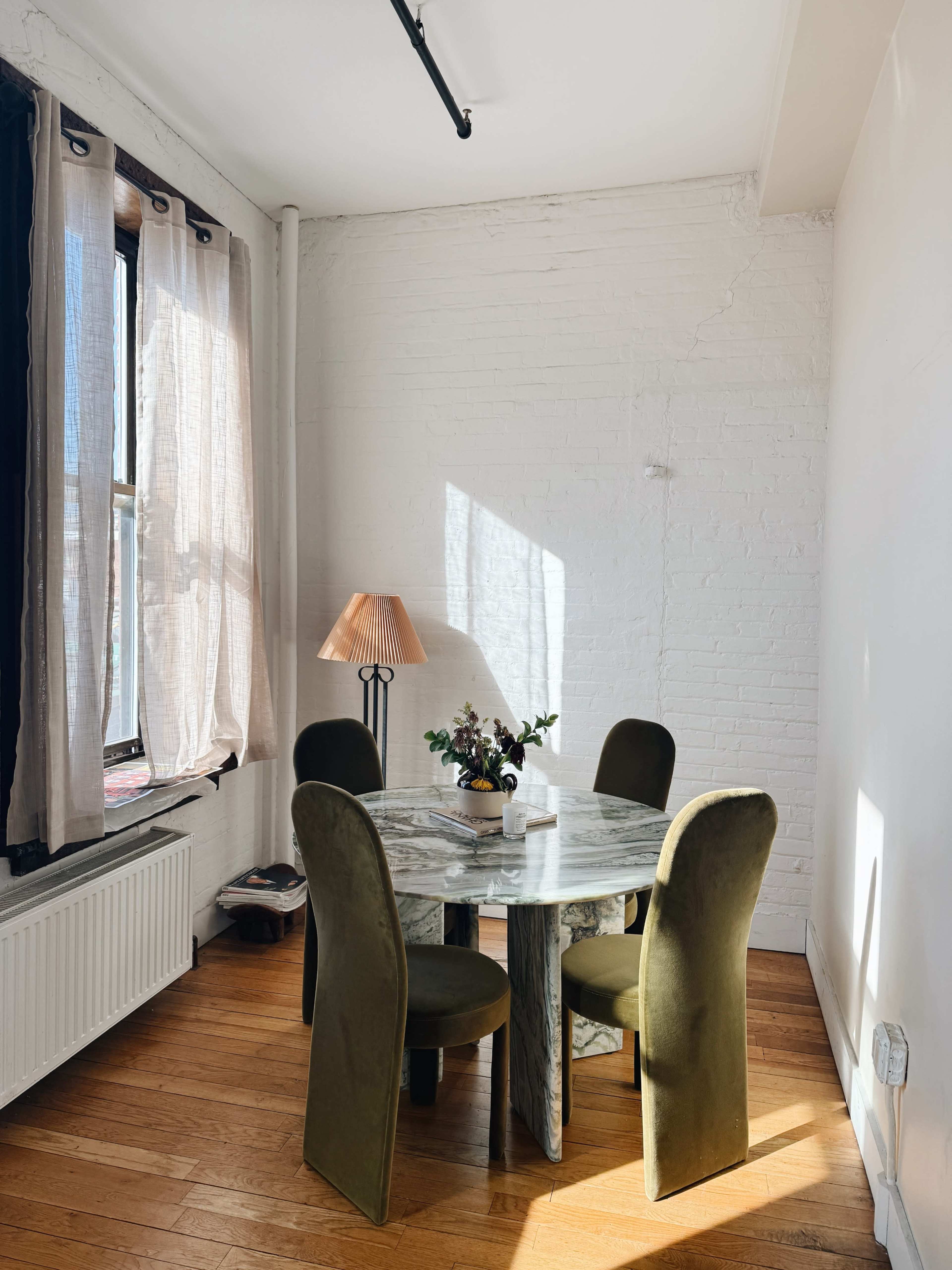 A dining area features a round marble table surrounded by four green upholstered chairs, illuminated by natural light coming through a window with sheer curtains.