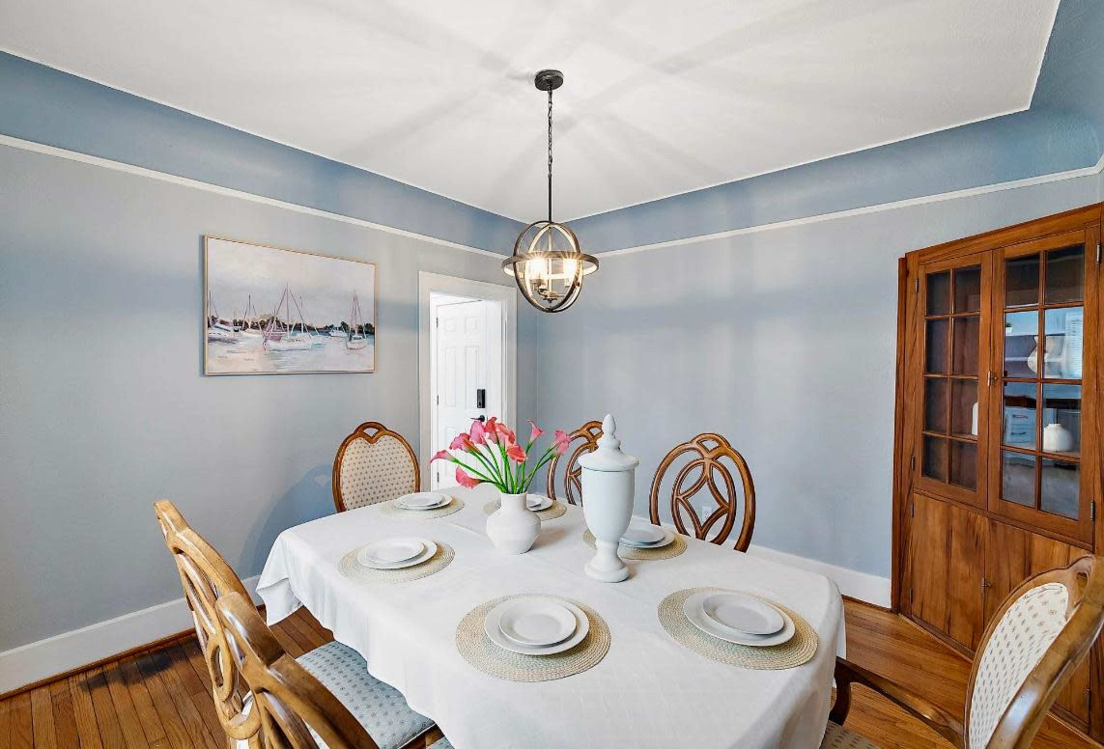 A dining room features a round table set for four, with a floral centerpiece and a painting of boats on the wall.